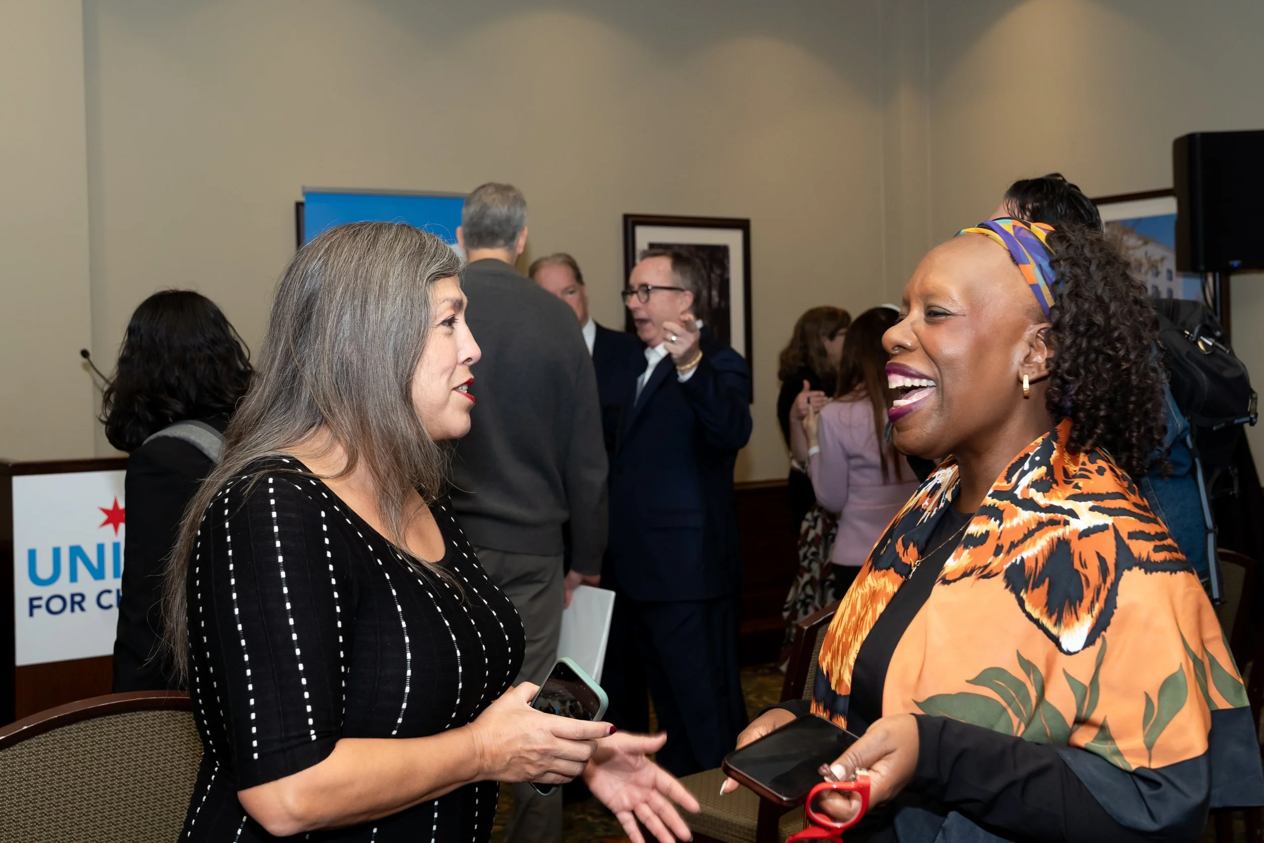 Two women are smiling and talking to each other at a crowded event, with other people in the background