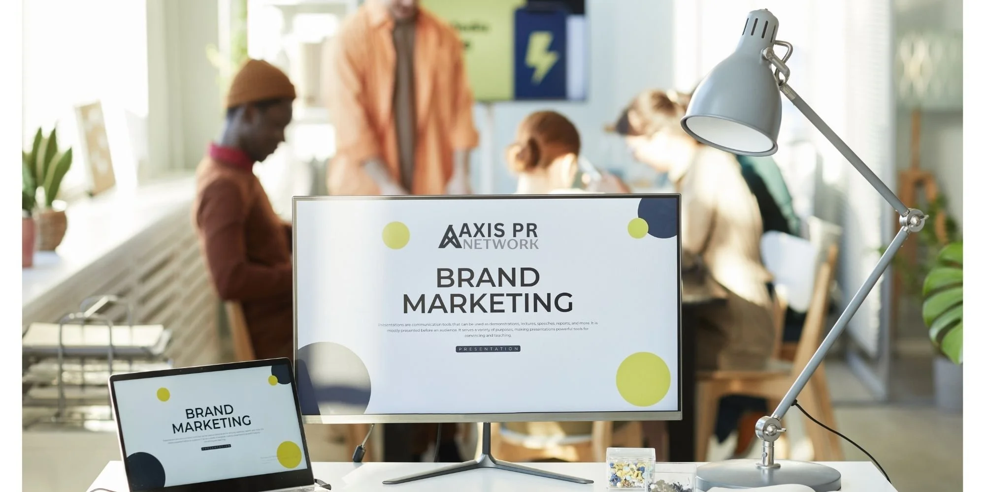 Office scene with a large monitor and a smaller laptop displaying 'Brand Marketing' for Axis PR Network, with a desk lamp and people in the background.