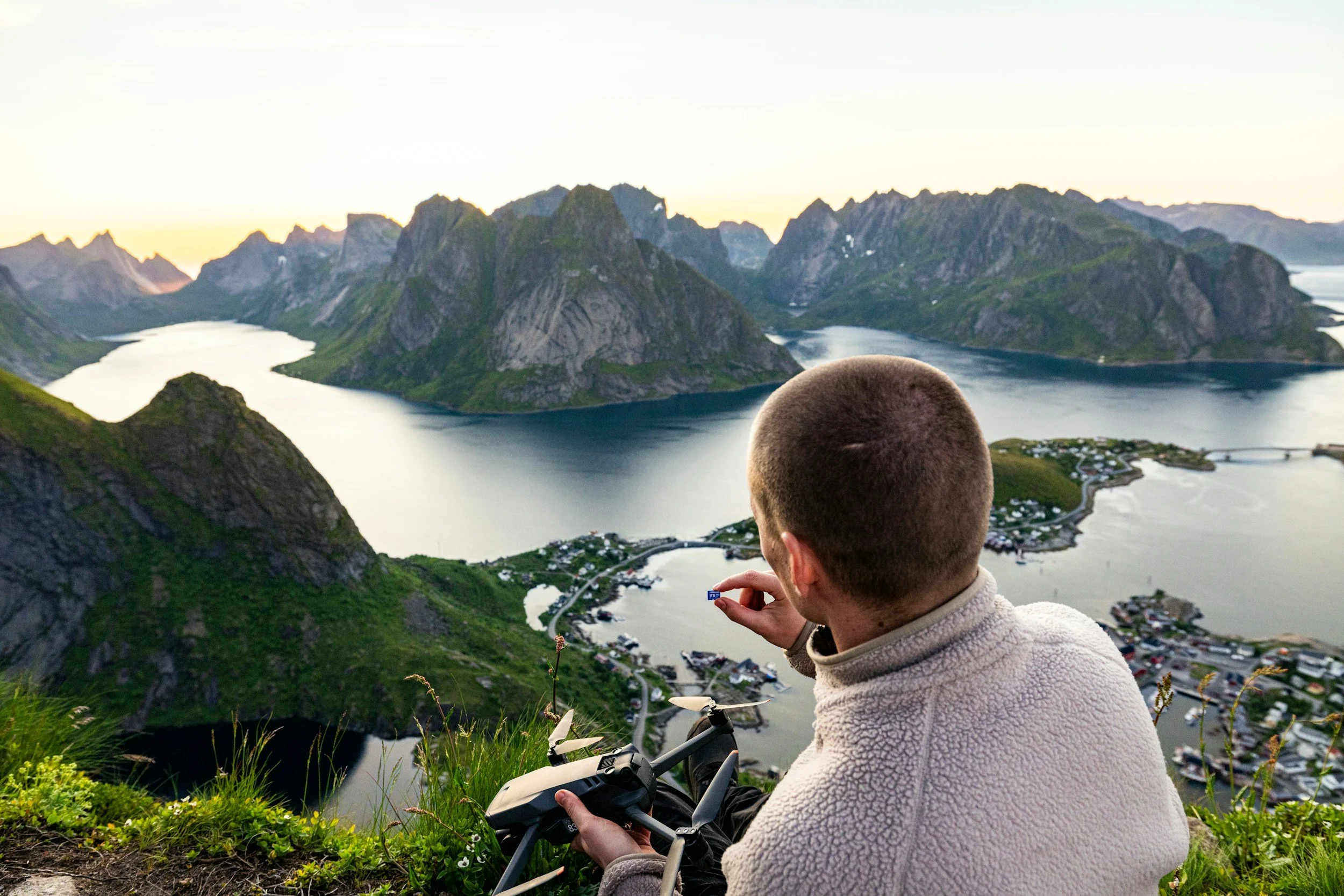 Man sitting on a grassy hill overlooking a fjord with mountains at sunset, holding a drone controller.