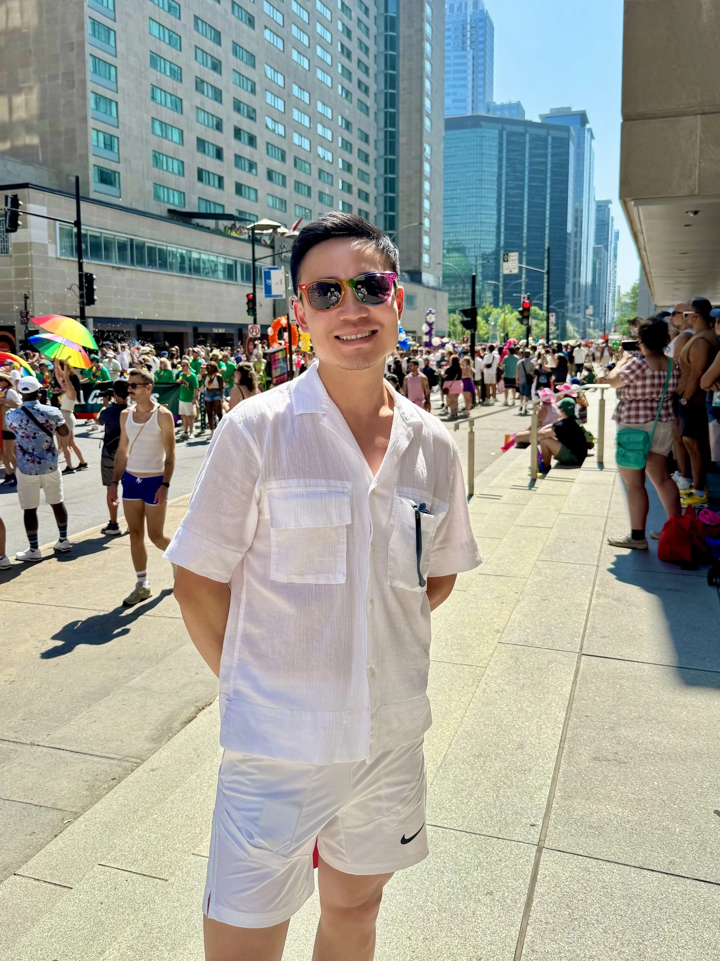 A man wearing sunglasses and a white button-up shirt stands with his hands behind his back in a crowded city street during a parade or celebration, with tall buildings and a diverse crowd in the background.