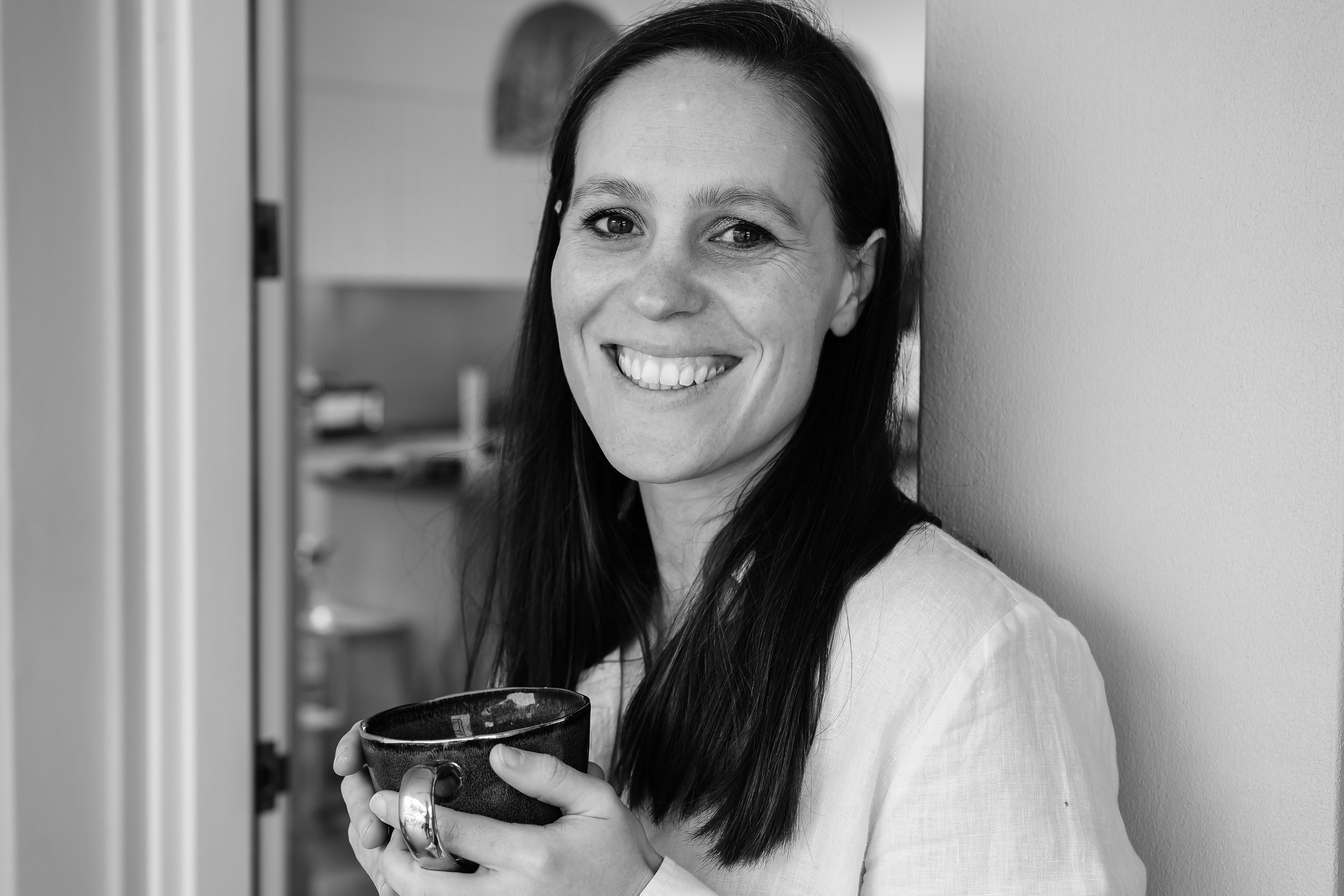 A woman with long dark hair smiling and holding a cup in her right hand, standing against a plain wall in an indoor setting.