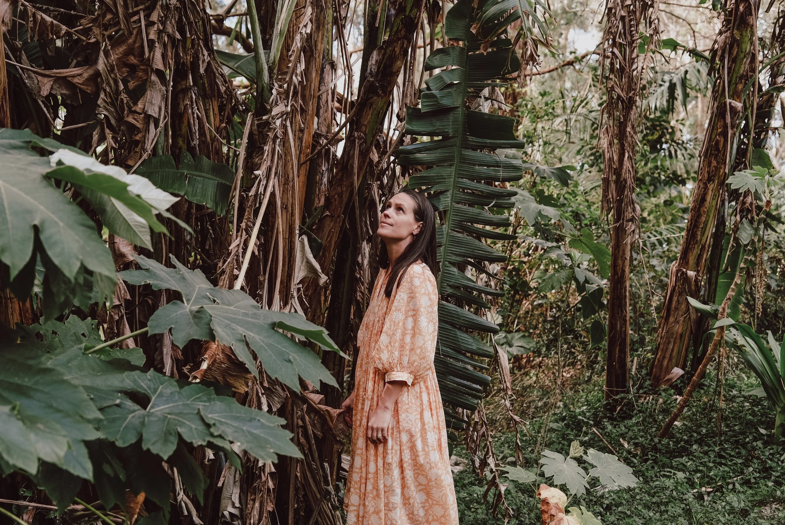 A woman with long dark hair in an orange floral dress standing among tall banana and other tropical plants in a lush jungle.