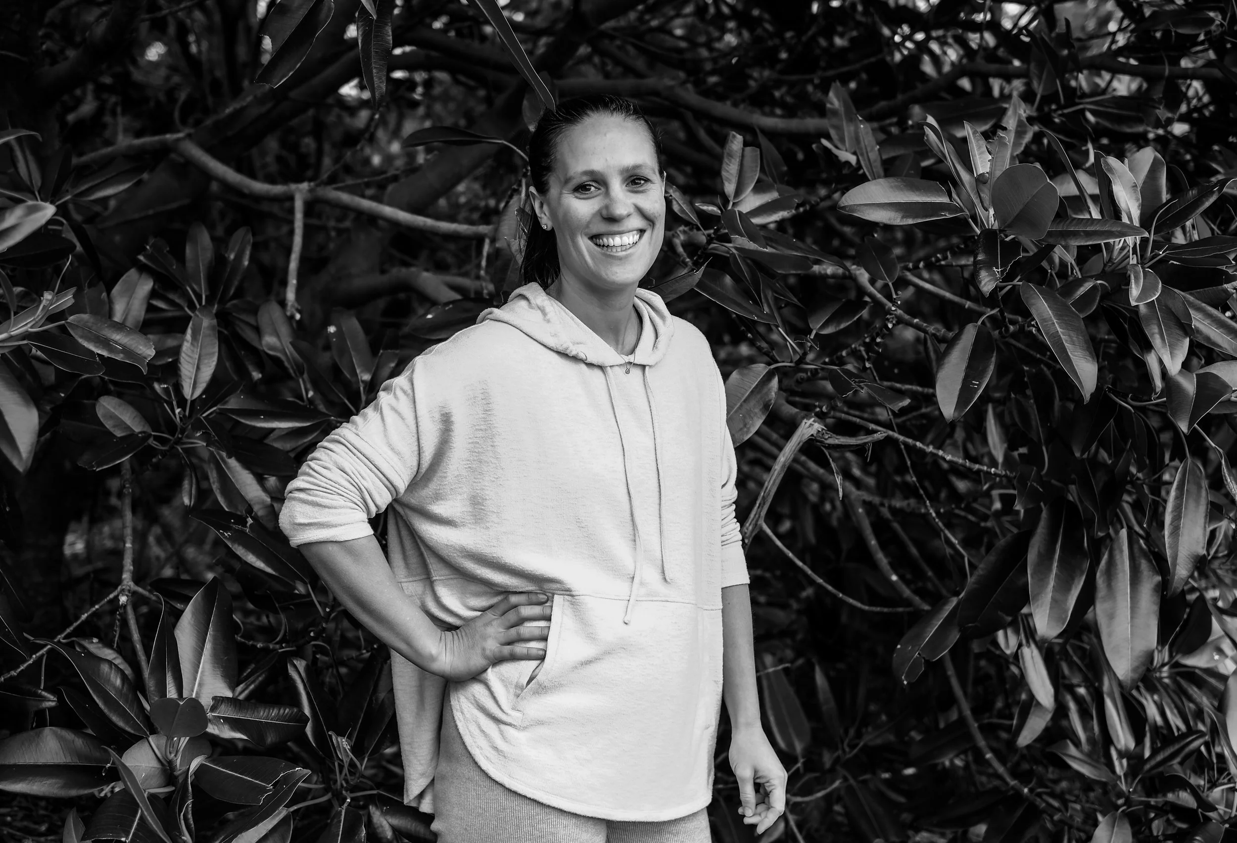A smiling woman standing in front of dense foliage with large leaves, wearing a hoodie and skirt.