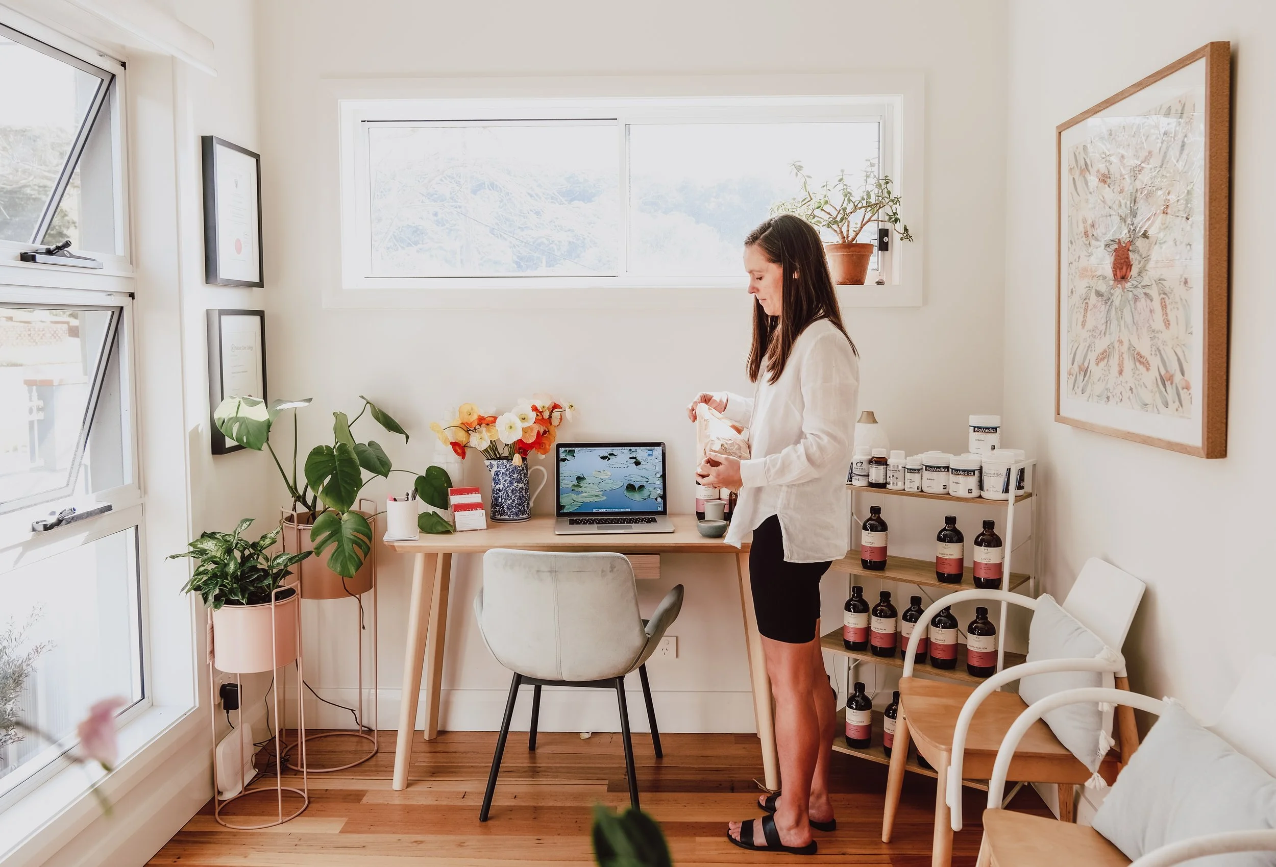 A woman with long dark hair, wearing a white jacket and black shorts, standing at a desk in a bright, cozy room. The room has white walls, a large window, various plants, framed artwork on the walls, a laptop on the desk, and shelves with bottles.
