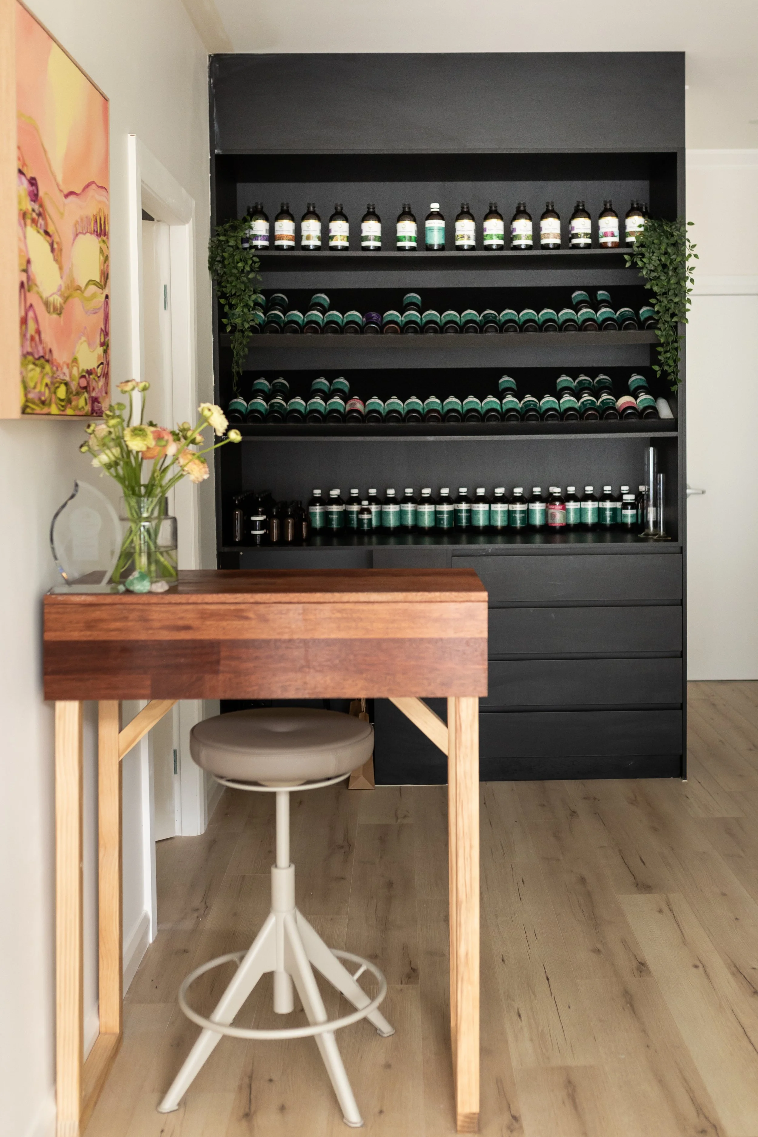 A black shelving unit filled with bottles and containers, flanked by green plants, in a room with white walls and light hardwood floors, seen from a side table with a flower vase and artwork on the wall.