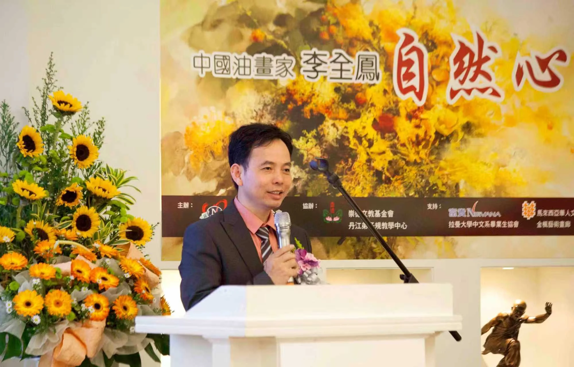 A man in a suit with a pink shirt and striped tie speaks into a microphone at a podium, positioned near a large floral arrangement of yellow sunflowers and orange blooms. Behind him is a colorful backdrop featuring Chinese characters and autumnal artwork, while a small bronze statue of a person in a dynamic pose is visible in the foreground.