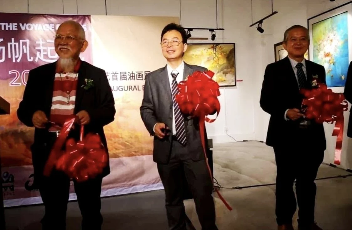 Three men at an art exhibition event, each holding a red ribbon and a small gift, standing in front of a backdrop featuring Chinese characters and landscape paintings on the wall.