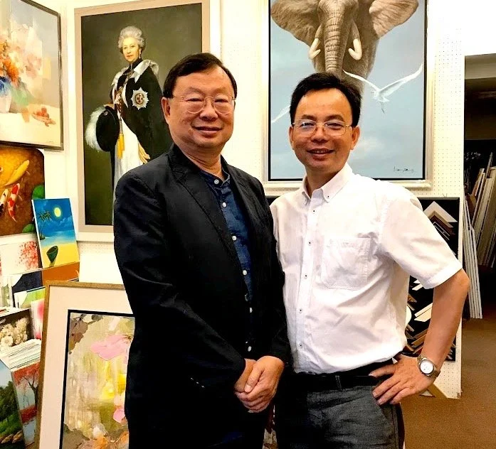 Two men stand together in an art gallery, smiling. Behind them are various paintings, including a portrait of a woman in historical attire, an elephant, and colorful abstract art.