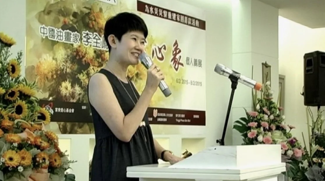A woman with short black hair singing into a microphone at an event with floral arrangements on both sides and a large banner in the background.