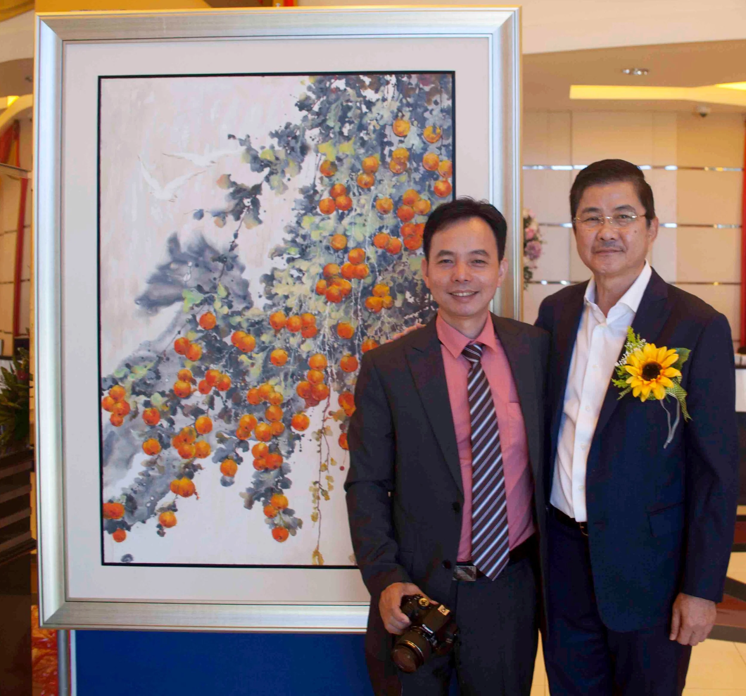 Two men in business suits stand before a painting of orange berries and green leaves, one holding a camera and the other wearing a sunflower boutonniere.