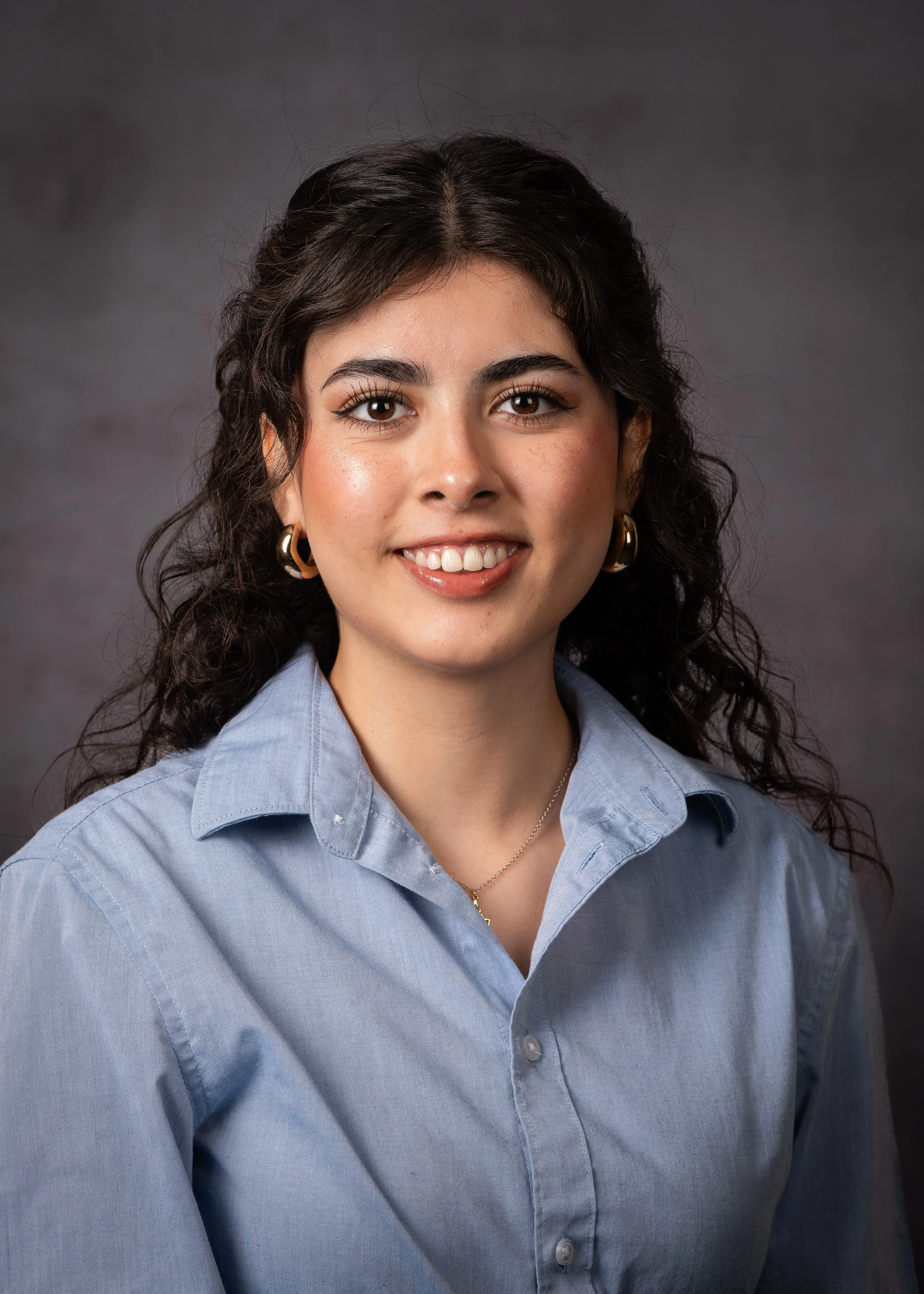 A young woman with dark curly hair, wearing gold hoop earrings and a light blue button-up shirt, smiling at the camera against a dark gray background.