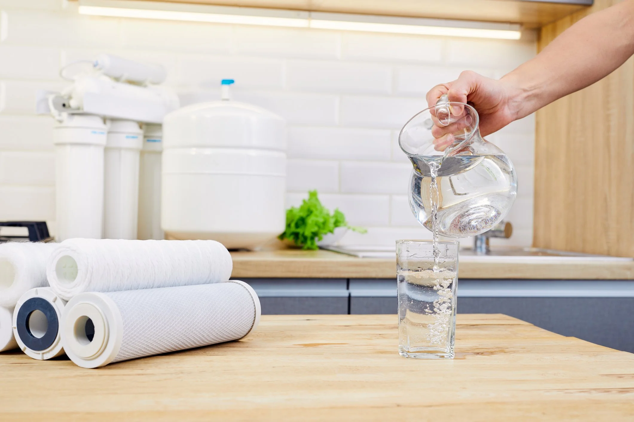 Person pouring water from a glass pitcher into a tall glass on a wooden kitchen countertop. Water is bubbling in the glass. Several water filter cartridges are on the countertop, with a white water filter system and a green plant in the background.