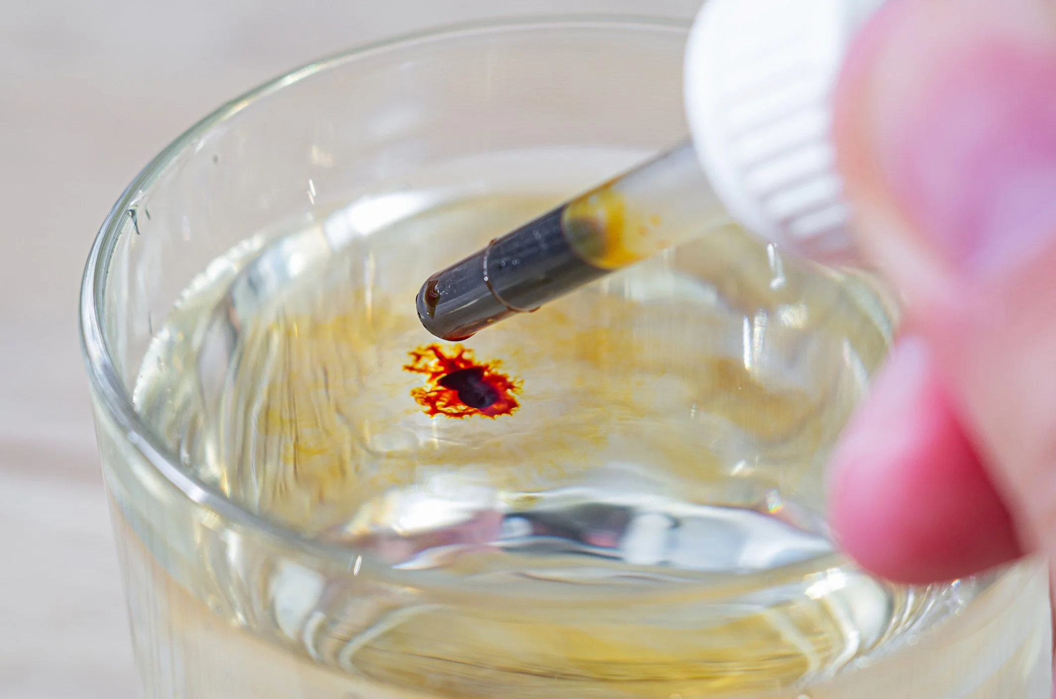 A close-up of a glass of water with a drop of red blood, with a hand holding a dropper above the glass.