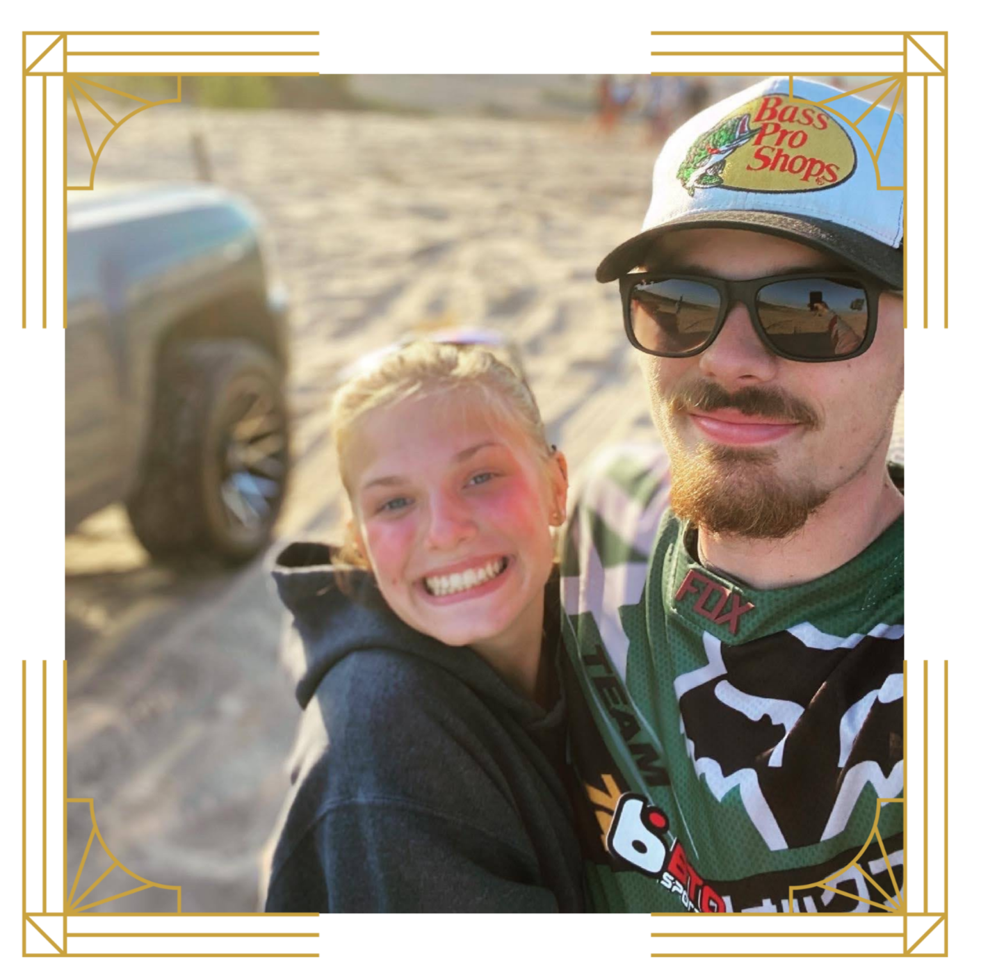 A smiling young woman and man taking a selfie at the beach. The man is wearing sunglasses, a baseball cap with a "Bass Pro Shops" logo, and motocross gear. The woman has blonde hair and is wearing a dark hoodie.