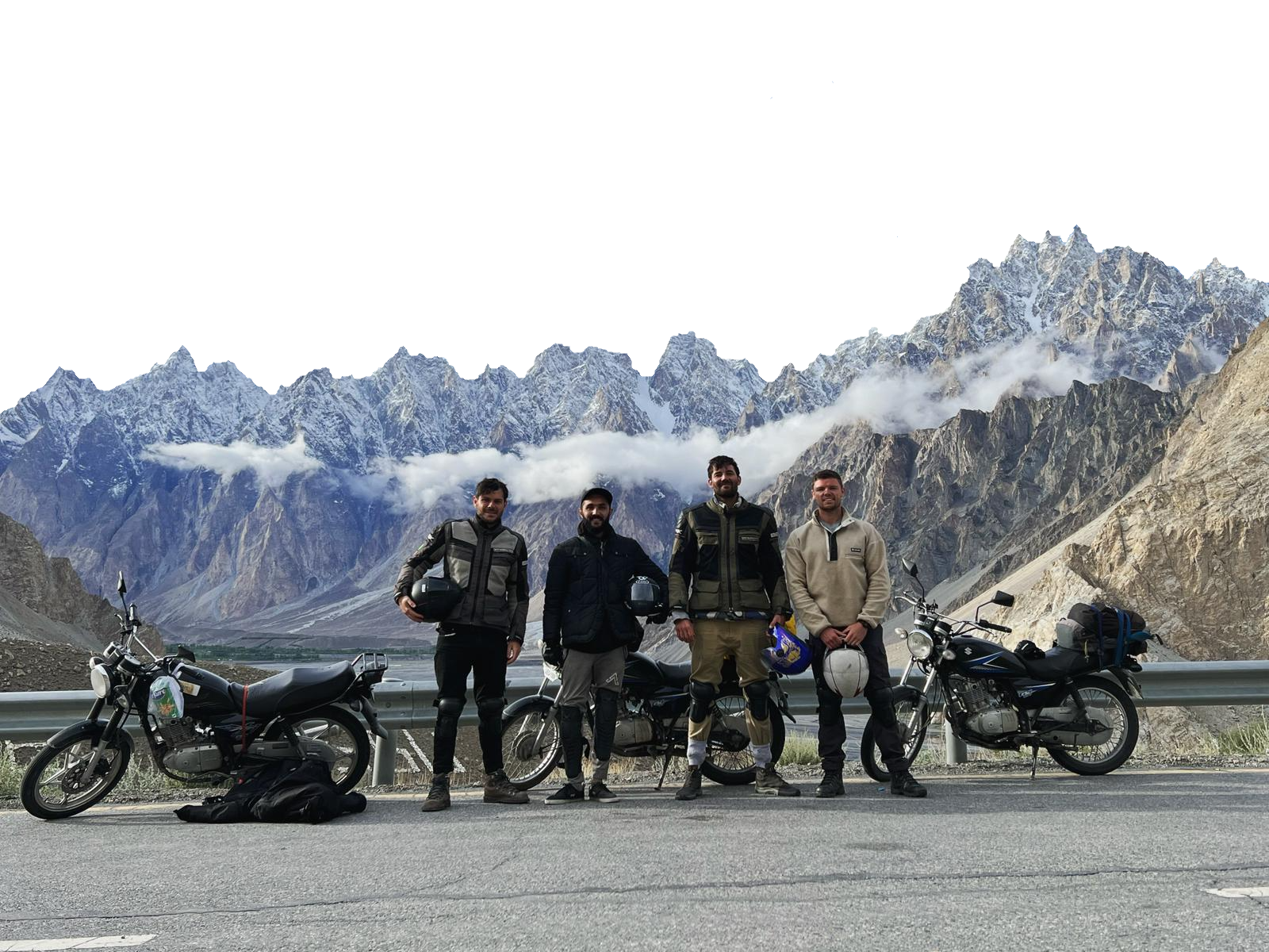 Four men in riding gear standing in front of their motorcycles on the Karakoram Highway with snow-capped peaks of Passu Cones and clouds in the background.