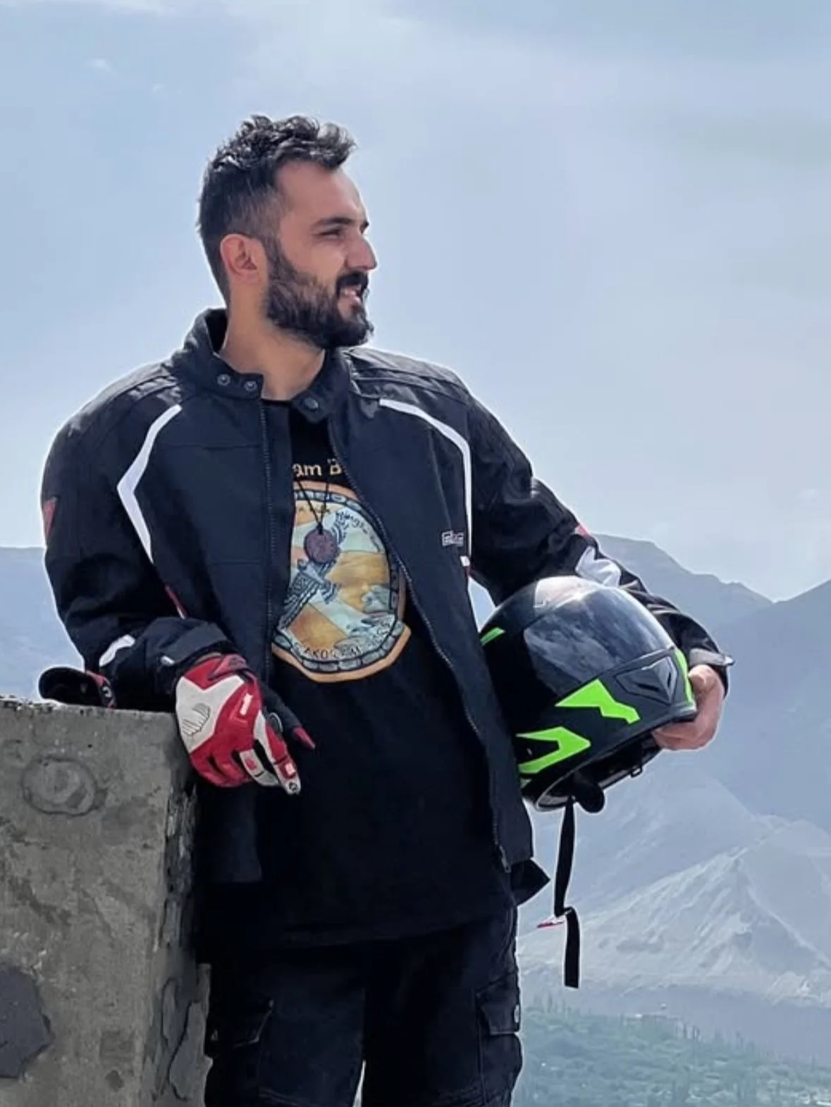 A man with a beard holding a black and neon green motorcycle helmet, wearing a black motorcycle jacket with white and red accents, standing outdoors with mountains and a cloudy sky in the background.