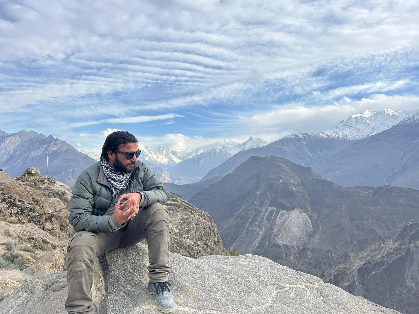 Man sitting on rock in mountains with snow-capped peaks, cloudy sky, wearing sunglasses, green jacket, and scarf. The location is Eagles Nest also known as Duikar lookout in Karimabad Hunza