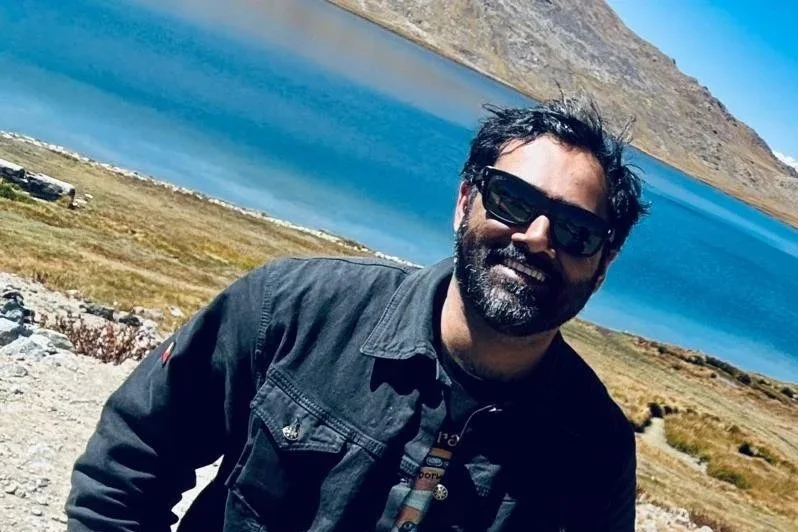 A man with sunglasses, a beard, and a black jacket smiling outdoors with a scenic background of a lake and mountainous landscape.