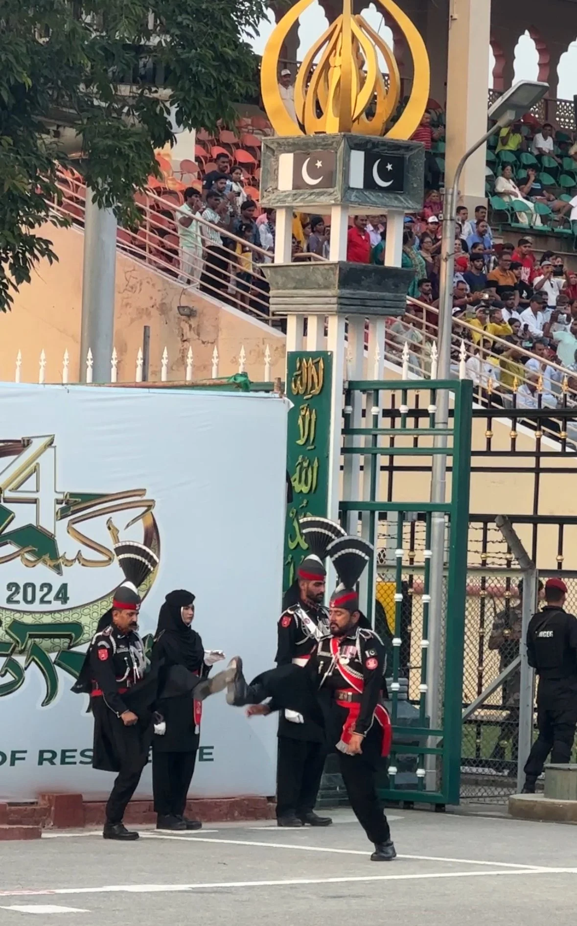 A ceremonial event with uniformed guards, one carrying another person. A large crowd is seated in a stadium in the background. Islamic symbols are visible on a sign and a lantern.