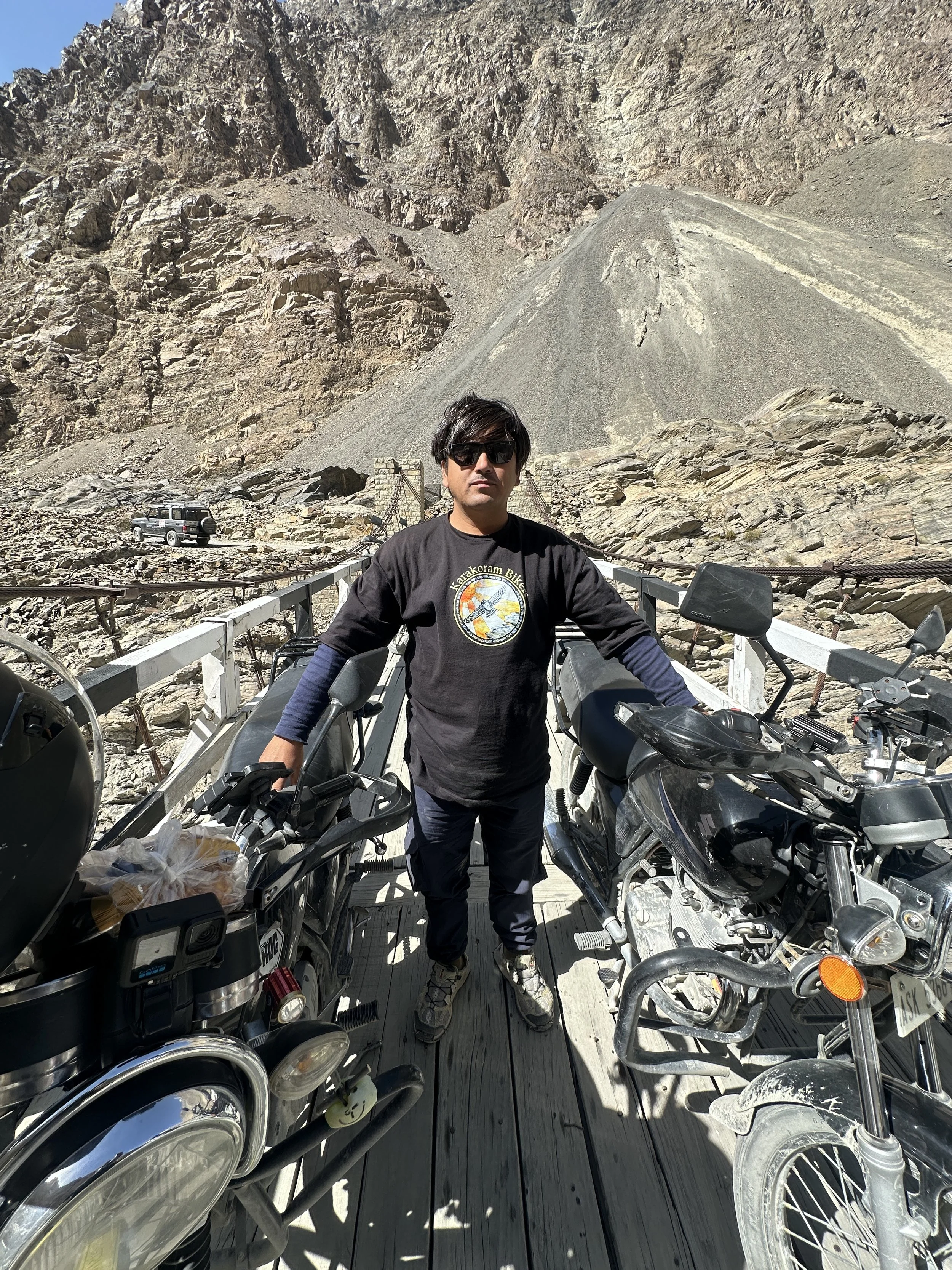 A man standing between two parked motorcycles on a wooden bridge in a mountainous area with rocky terrain and steep slopes in the background.
