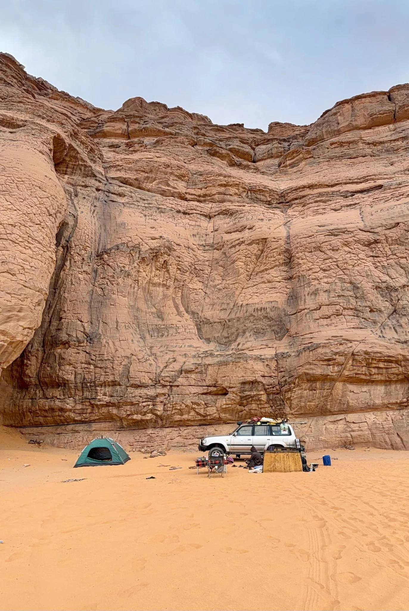 Camping setup in a desert with a tent, a white off-road vehicle, and camping gear against a towering sandstone cliff. Algeria motorcycle tour. KB Global motorcyle tours. Karakoram Bikers Algeria