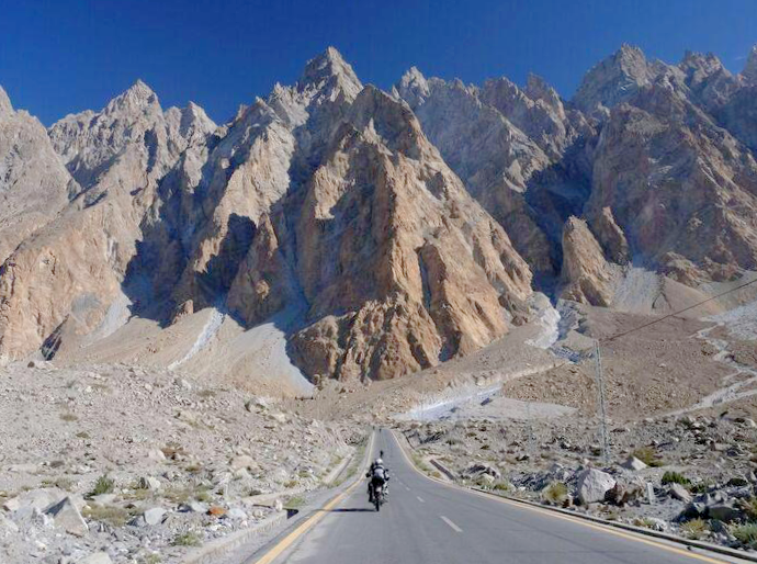 A motorcyclist with Karakoram Bikers adventure tours riding on the Karakoram Highway near passu surrounded by rocky, rugged mountain peaks under a clear blue sky.