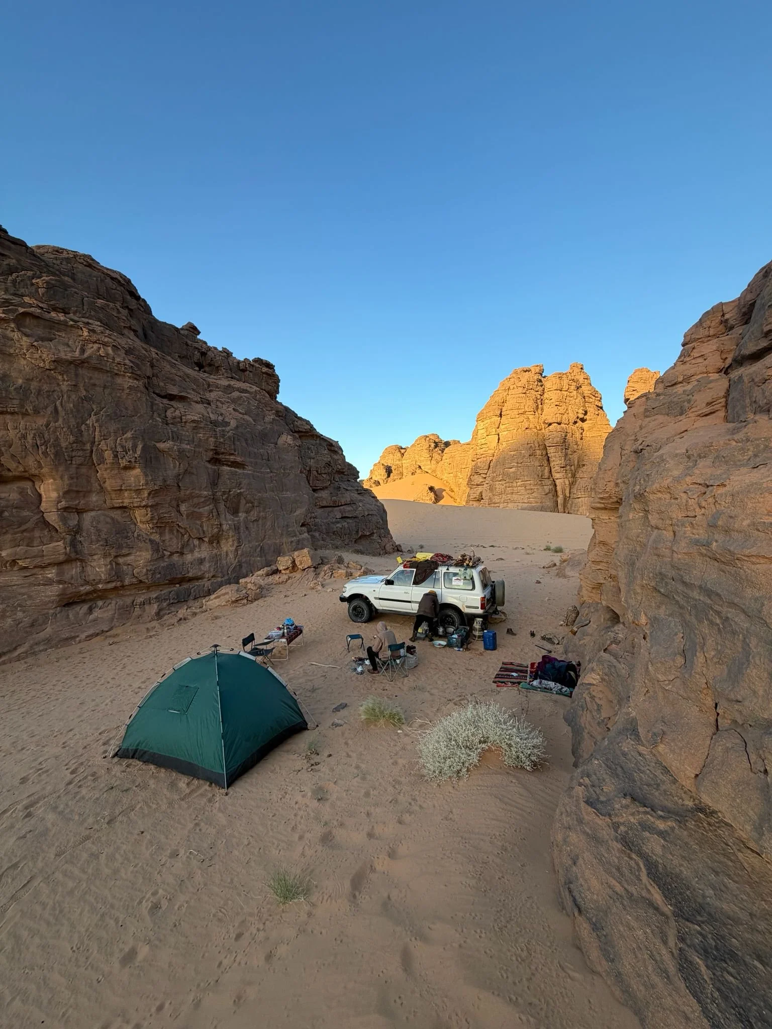 A desert campsite with a green tent and a white vehicle parked among large rock formations. The sky is clear and blue with sunlight illuminating the rocks in the background. Algeria motorcycle tour. KB Global motorcyle tours. Karakoram Bikers Algeria