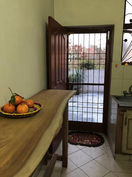A kitchen with a wooden table holding a decorative basket of oranges, a door with bars leading outside, and part of a sink and cabinet on the right.