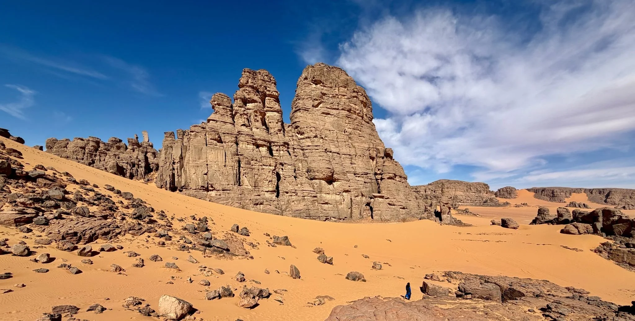 Tall rock formations in a desert with sandy dunes, scattered rocks, and a person walking. Clear blue sky with some clouds. Algeria motorcycle tour. KB Global motorcyle tours. Karakoram Bikers Algeria