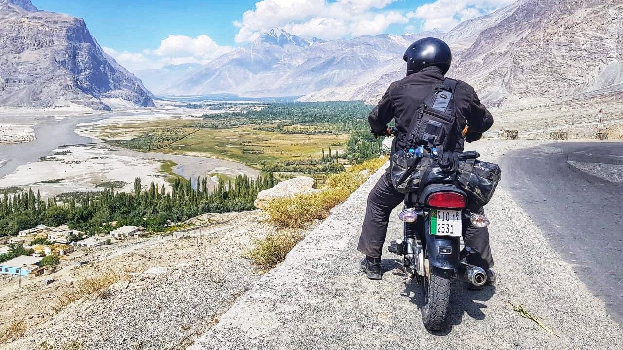 Motorcyclist wearing black biking gear and helmet riding on a mountain road overlooking a green valley with trees, a river, and mountains in the background. The Valley is Shigar and the motorbike is from Karakoram Bikers adventure tours in Pakistan.