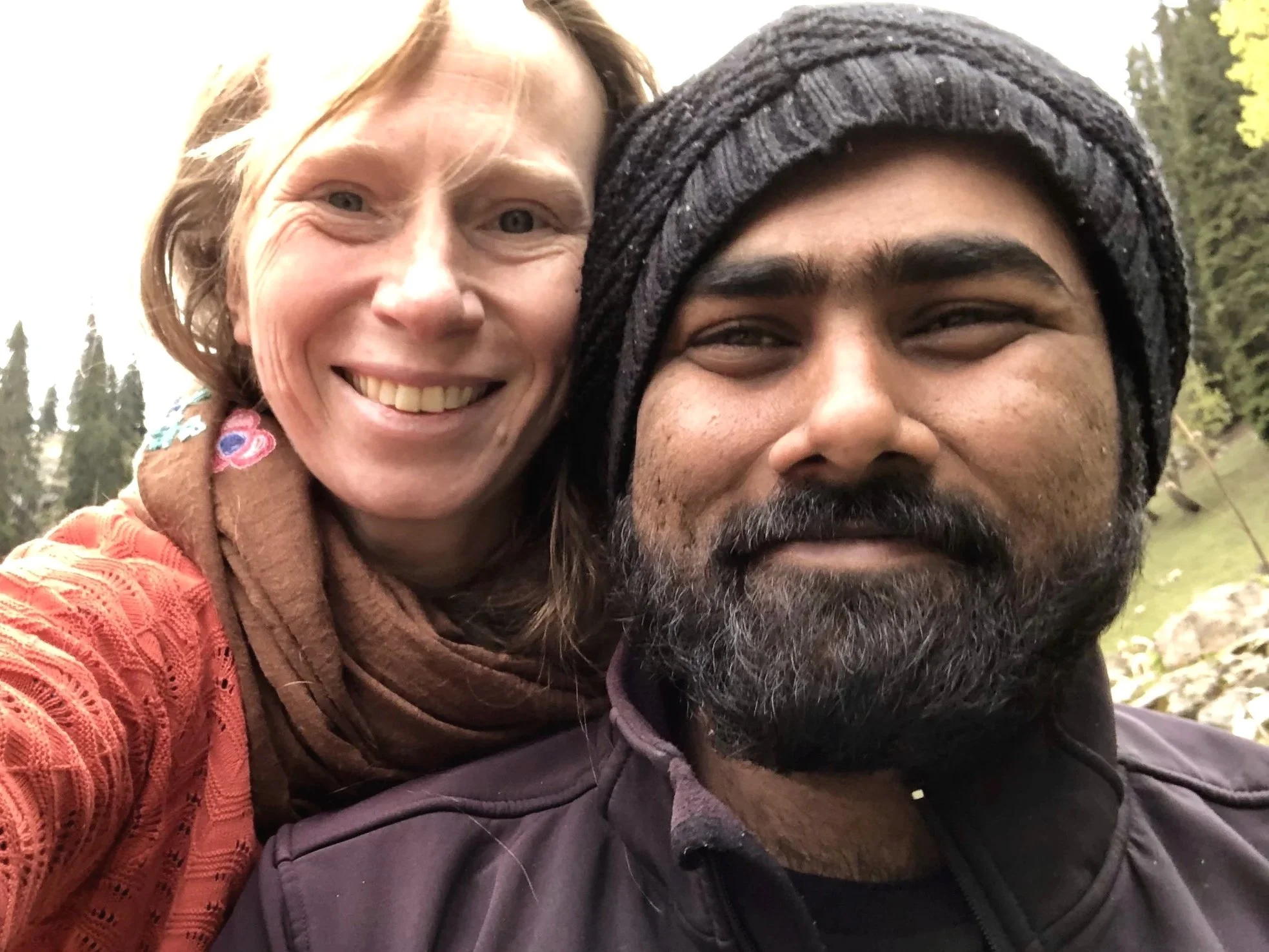 Close-up of a smiling woman with red hair and a brown scarf, next to a man with a beard, mustache, and wearing a gray knit hat outdoors with trees in the background.