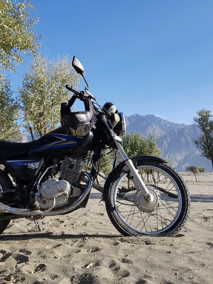 A black motorcycle parked on sandy terrain with trees and mountains in the background and a clear blue sky.