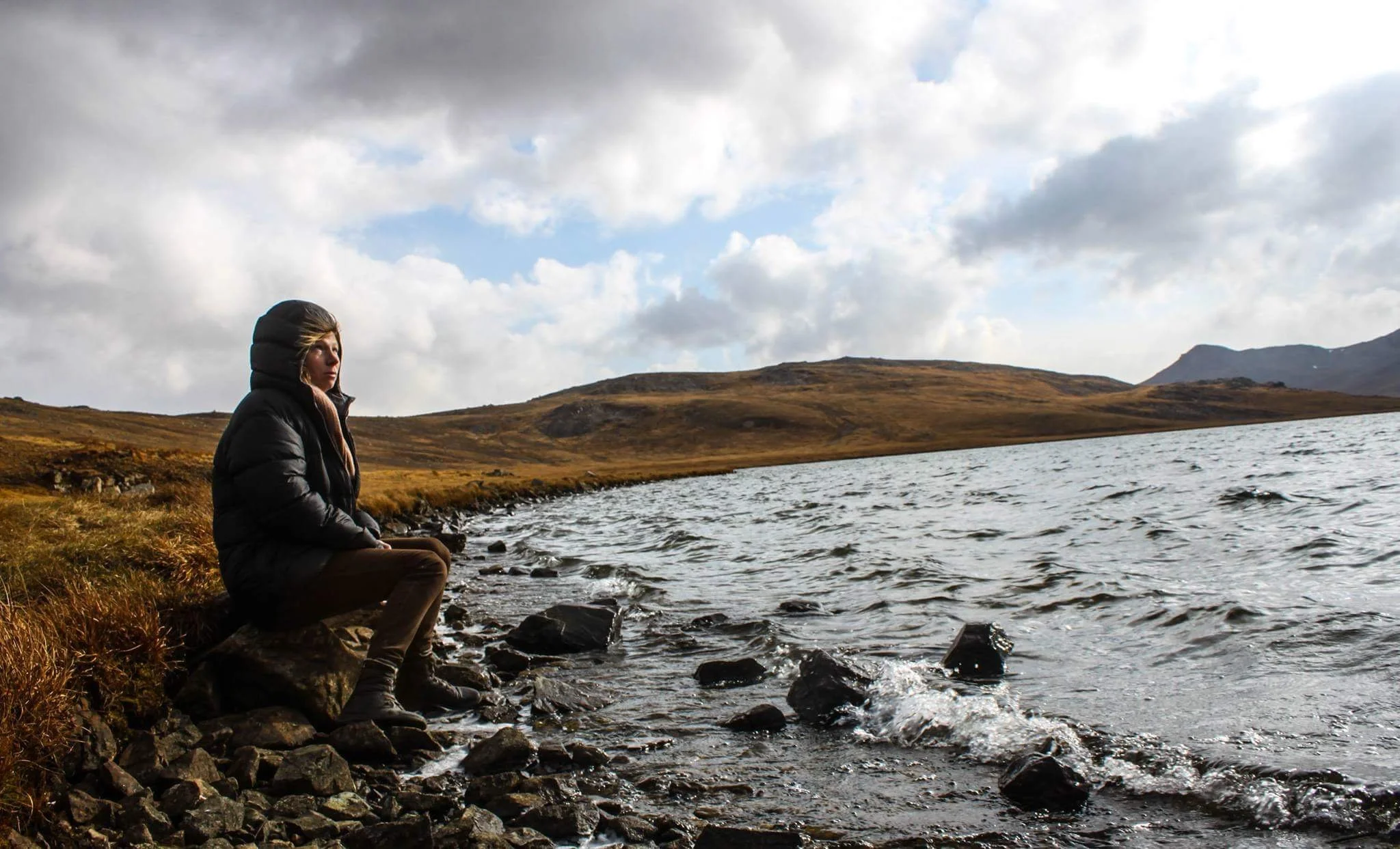 A woman sitting on rocks by a lake in a hilly landscape during cloudy weather. The lake is called Sheosar lake at Deosai National Park in Pakistan.