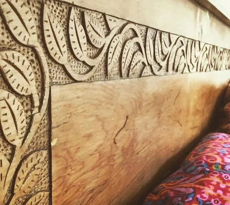 Close-up of a wooden headboard with carved leaf patterns and a colorful blanket on a bed.