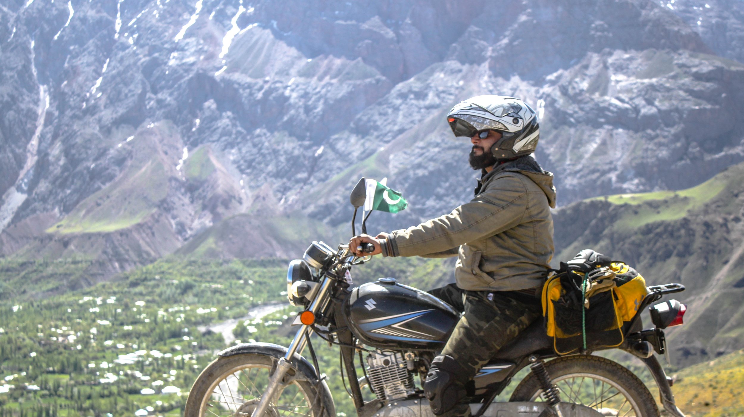 Man wearing helmet and sunglasses riding a motorcycle from Karakoram Bikers motorcycle tours in pakistan with a Pakistani flag on the mirror, with mountains and a valley in the background.