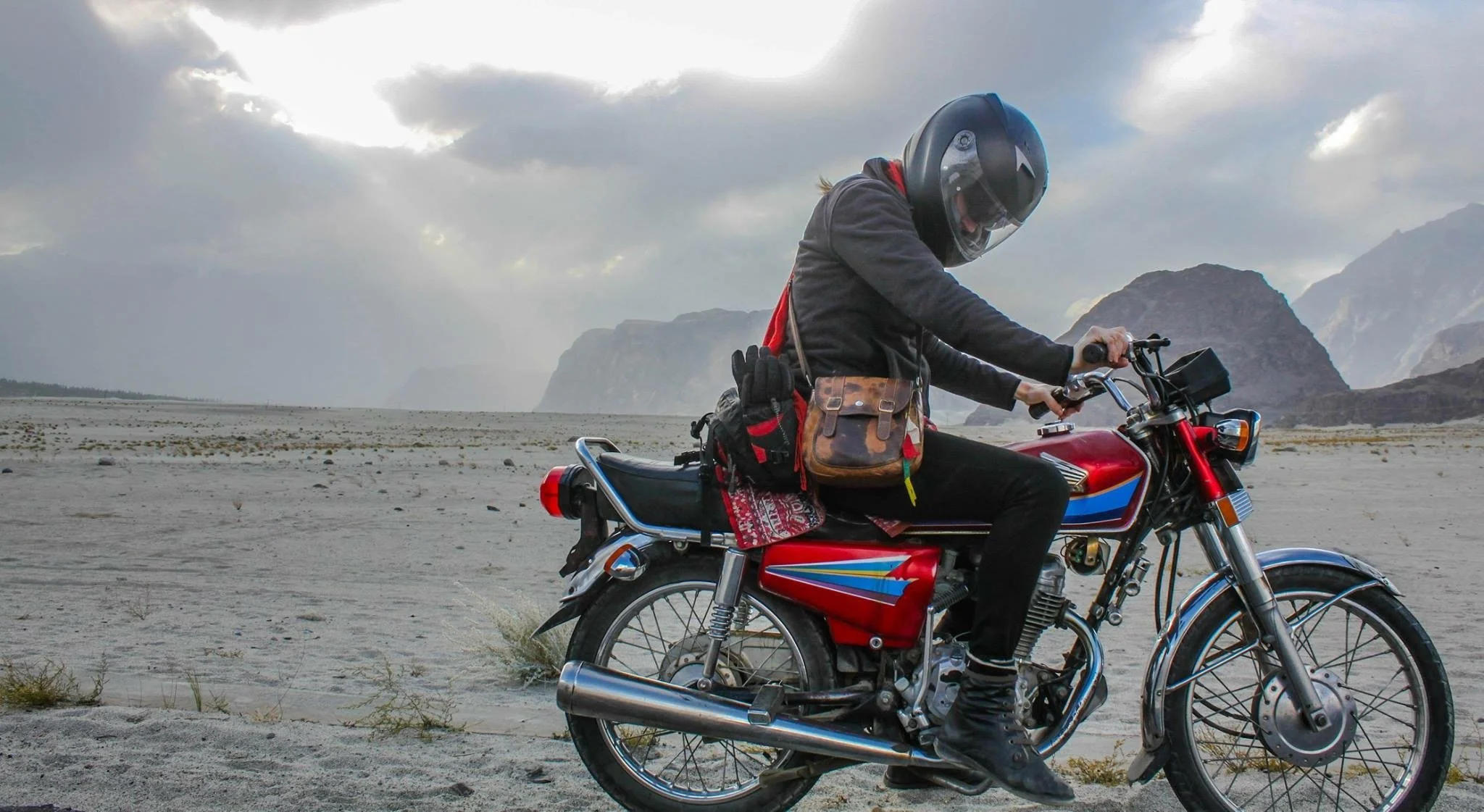 Person riding a red motorcycle from Karakoram Bikers adventure tour company on a sandy, desert-like landscape with mountains in the background, wearing a helmet, black jacket, and carrying a brown bag and gloves.