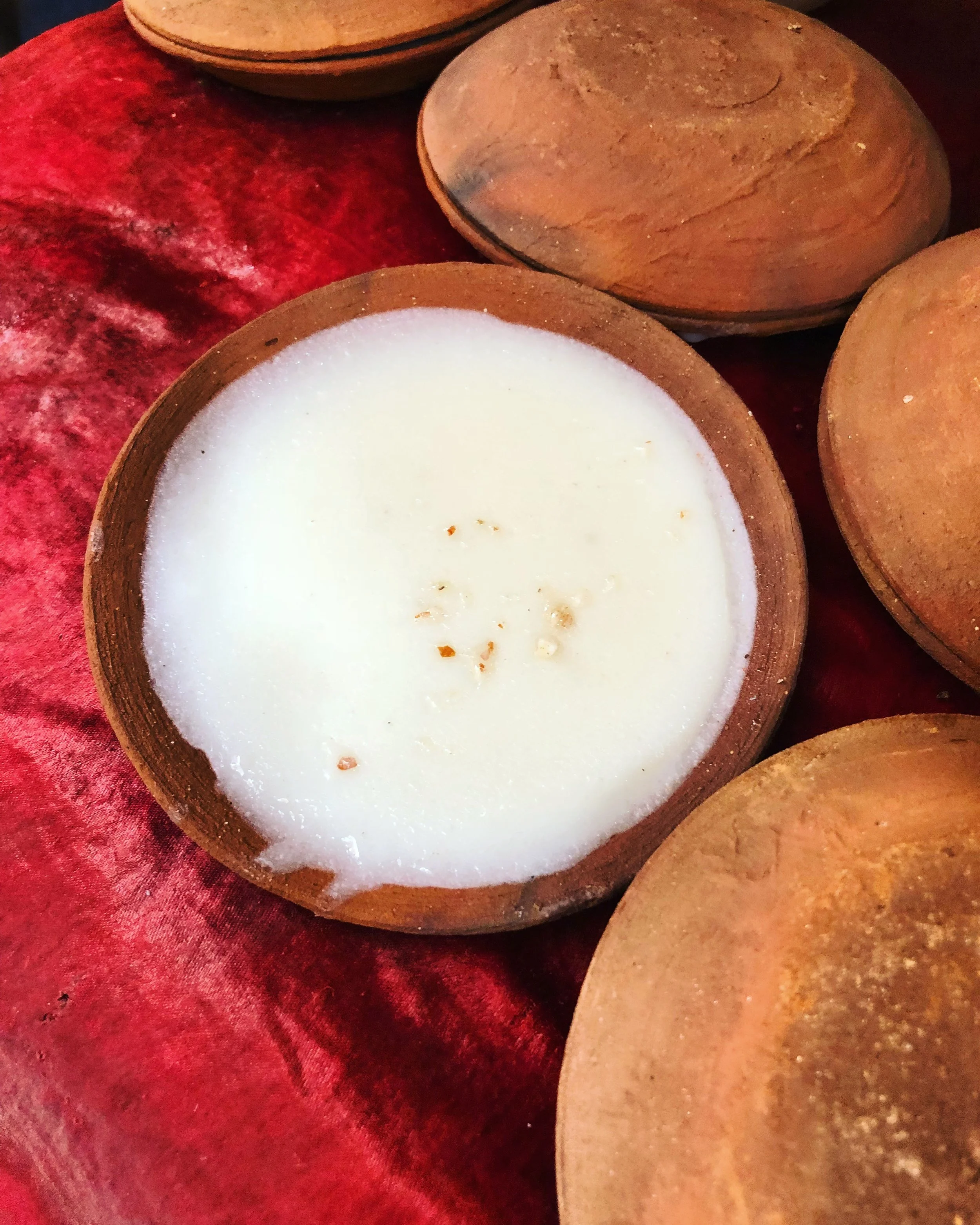 A wooden bowl filled with a white, foamy substance, possibly a traditional handmade soap or curd, surrounded by other wooden bowls on a red fabric surface.