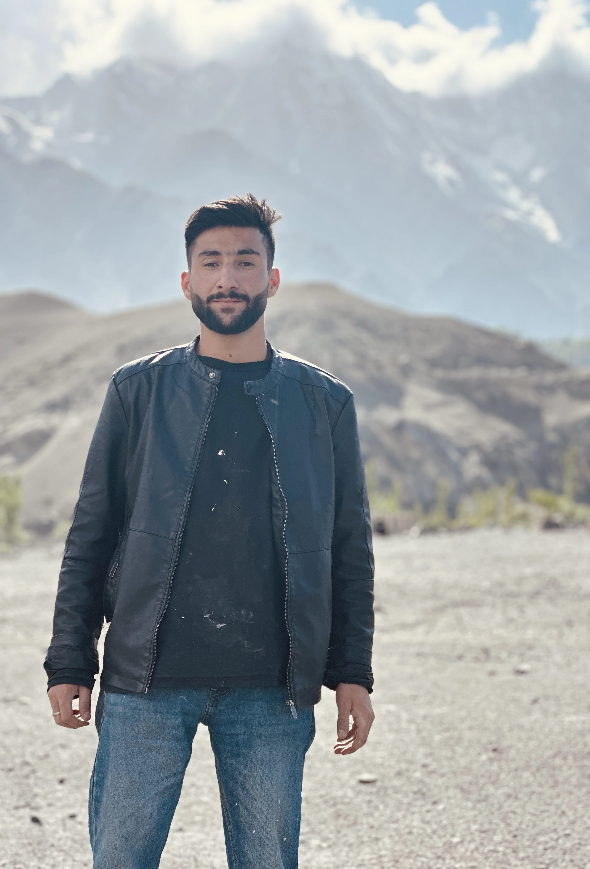 A man with dark hair and a beard stands outdoors in front of a mountain range with snow-capped peaks and a cloudy sky, wearing a black leather jacket and jeans.