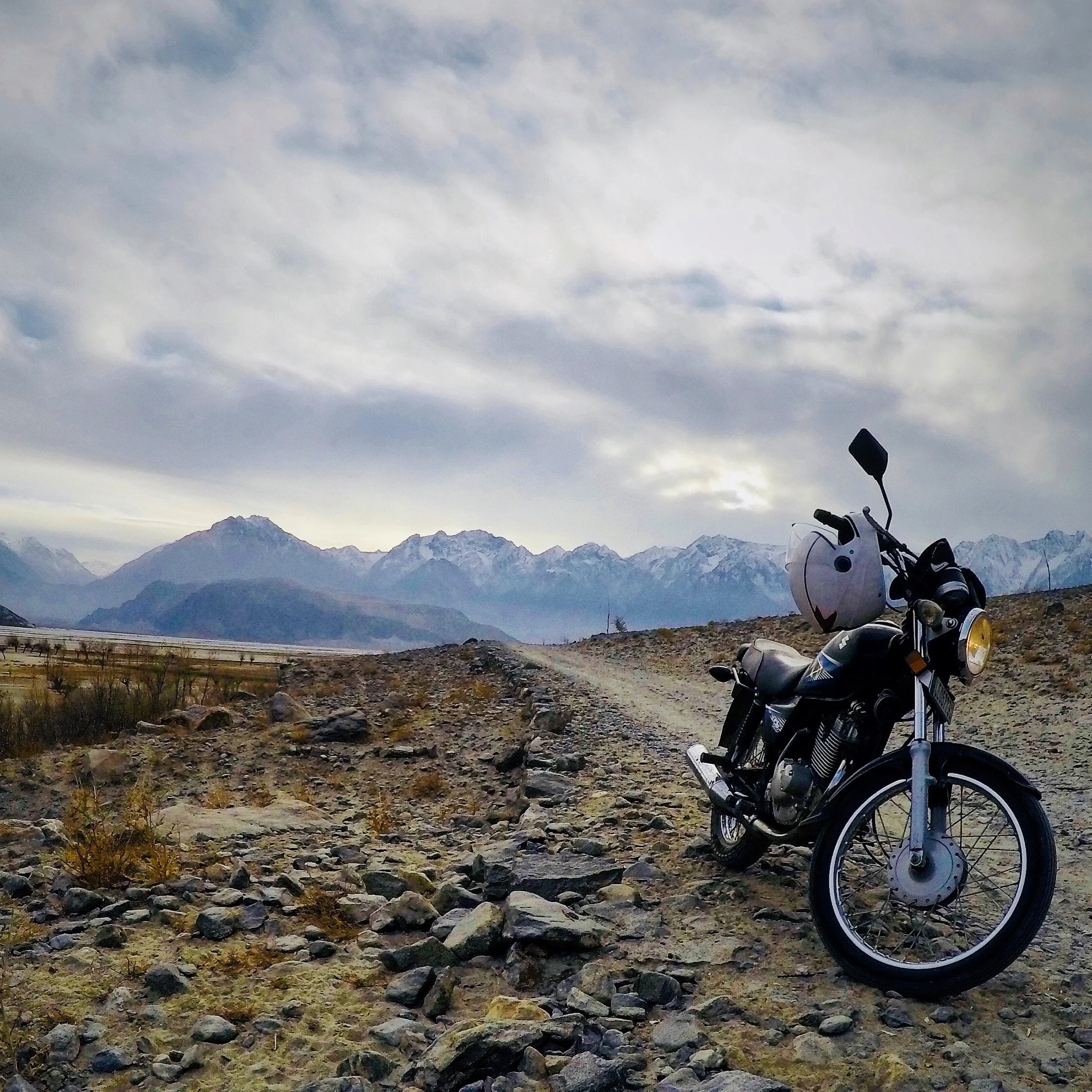 A motorcycle parked on a rocky dirt road in a mountainous landscape with snow-capped peaks and cloudy sky in the background.