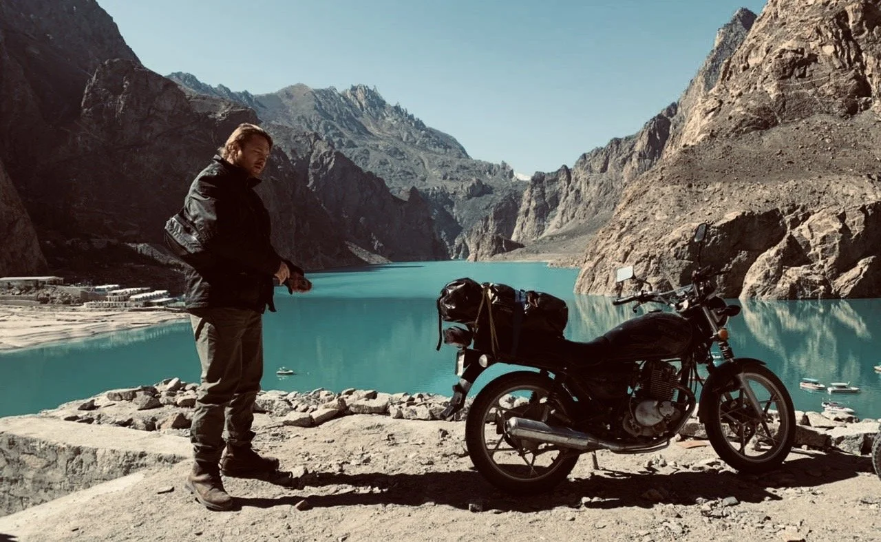 A man standing next to a motorcycle with a mountain lake and rugged cliffs in the background.