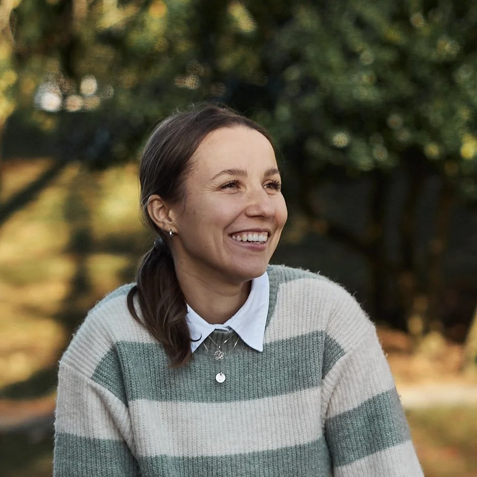 A woman smiling outdoors with trees in the background.