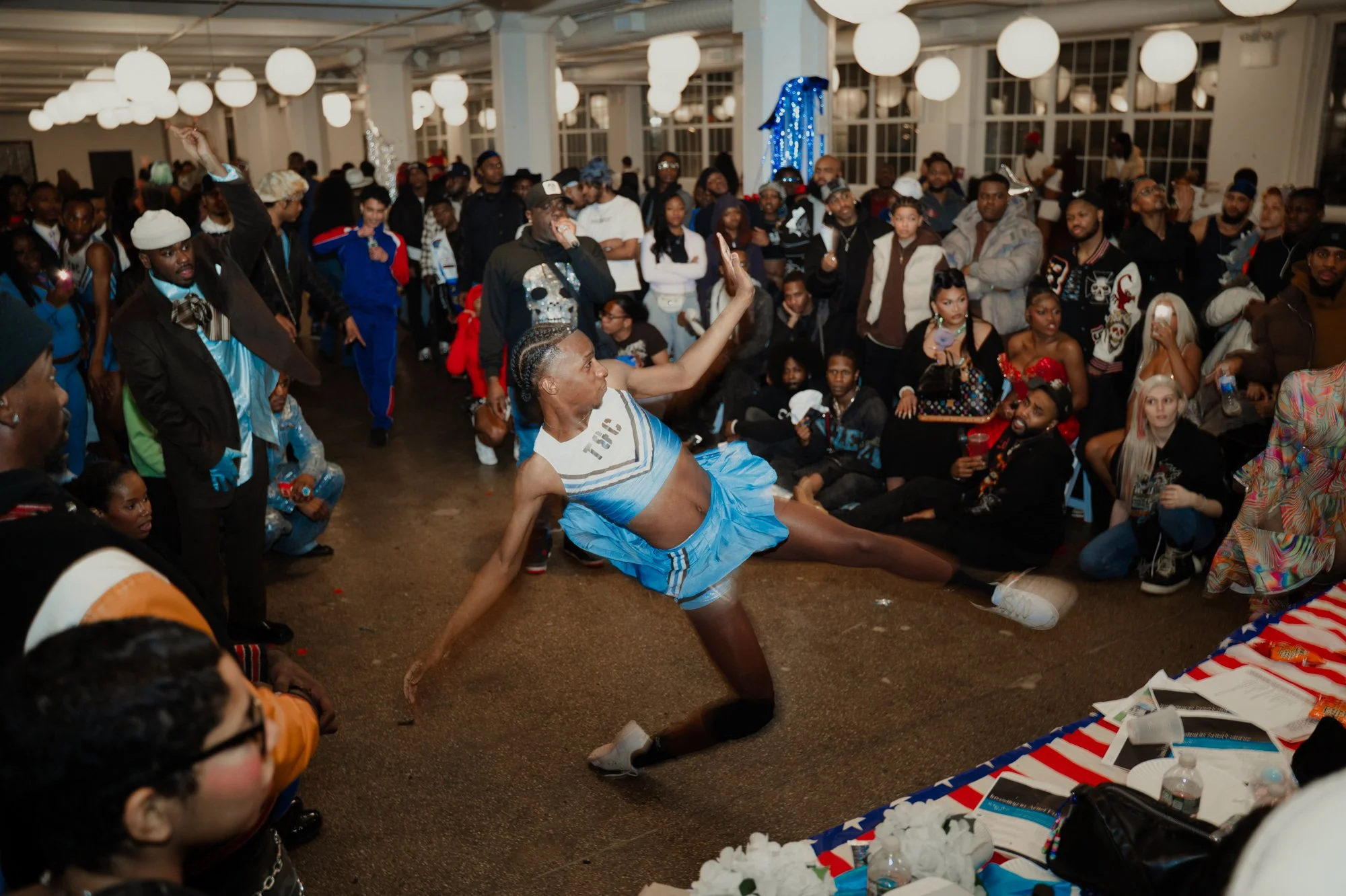 A dancer in a blue skirt performs a split on the floor at a crowded indoor event with many onlookers, balloons, and decorations.