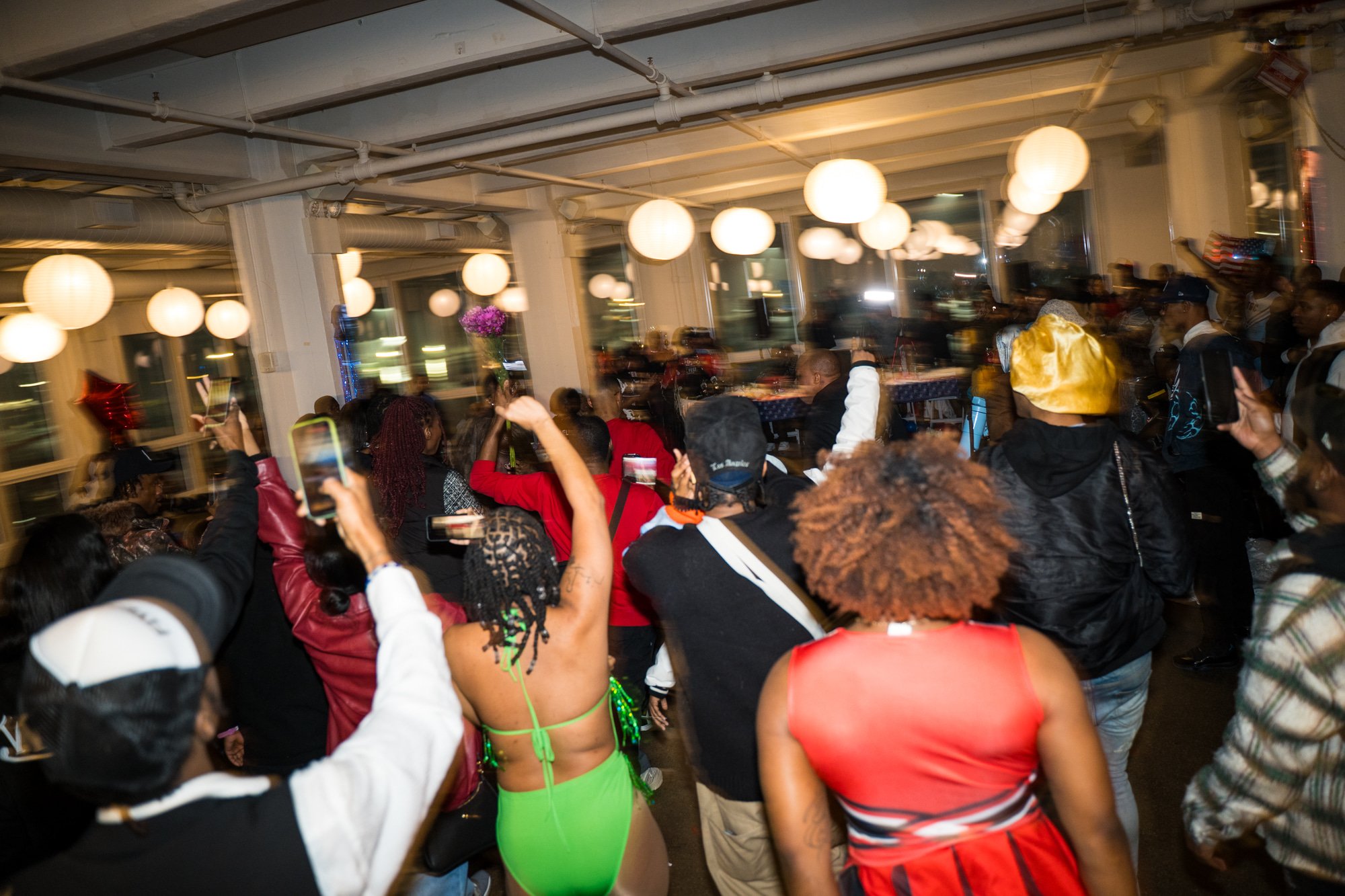 People gathered at an indoor event, many holding up phones and taking pictures. The room is decorated with hanging white lanterns, and there are balloons and a flower arrangement visible. The attendees are dressed in a variety of clothing styles, some casual and some more party-oriented.