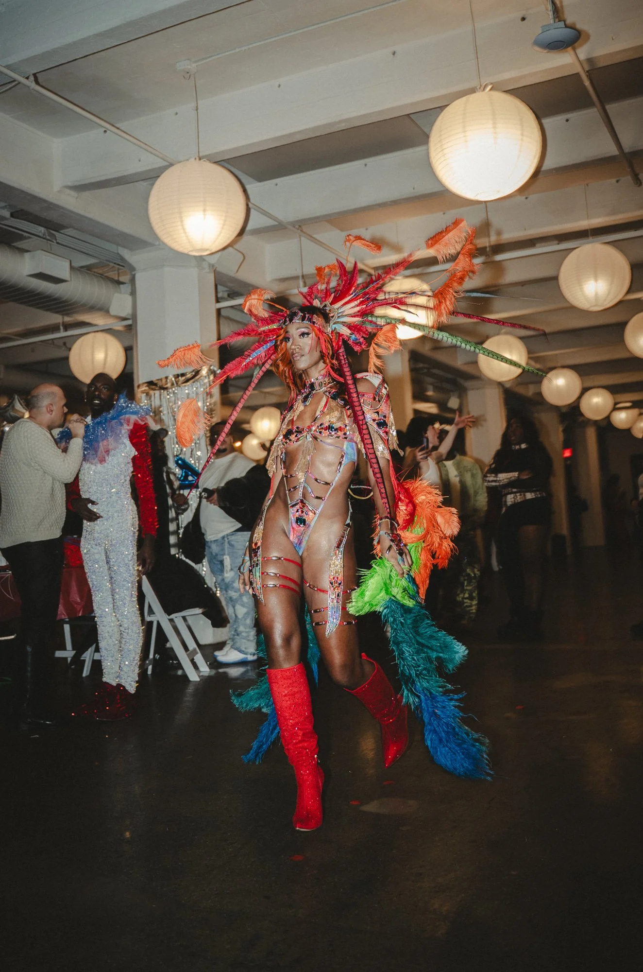 A person dressed in a colorful, feathered costume with red, orange, green, and blue feathers, including a large feathered headdress, red high-heeled boots, and revealing clothing, at a party or event with other people in the background.
