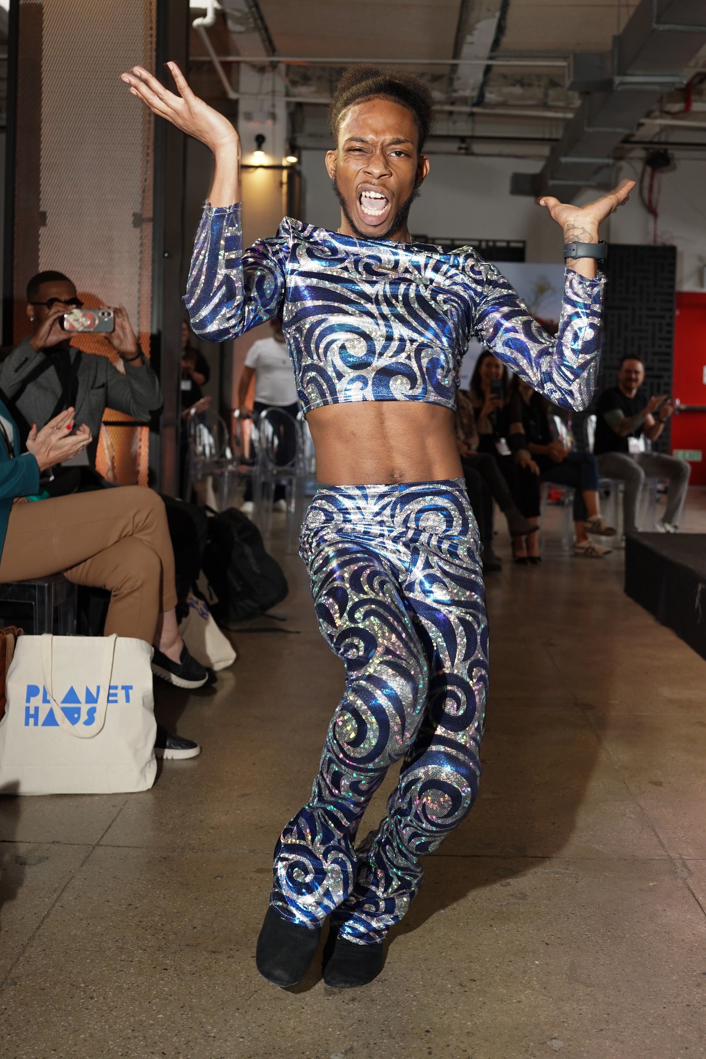 Person dancing in a shiny, swirling blue and silver outfit with a crop top and matching pants, performing energetically at an indoor event with seated audience members watching and taking photos.