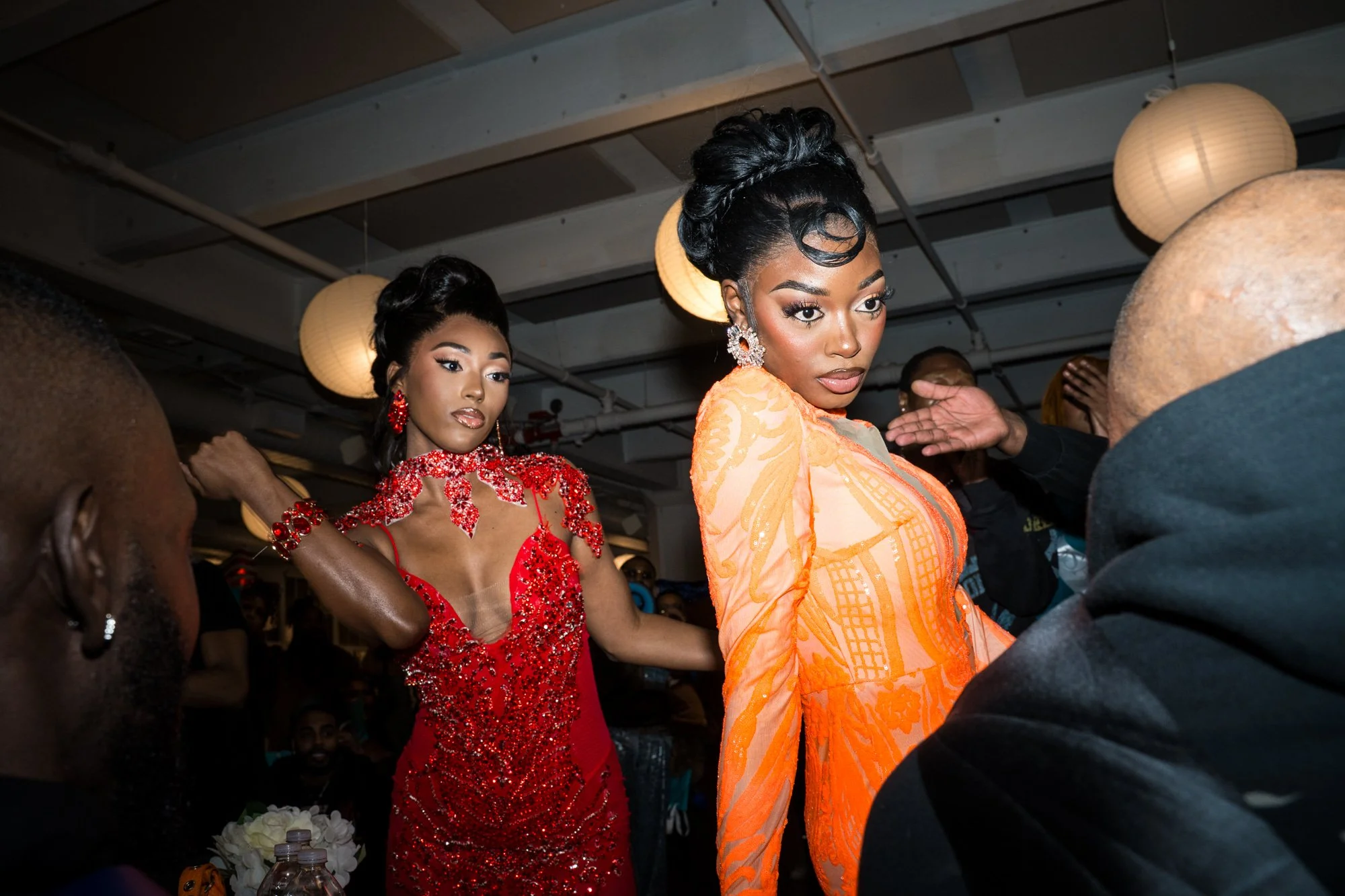 Two women dressed in vibrant, ornate clothing with elaborate hairstyles and jewelry, possibly at a fashion event or cultural celebration, standing among a crowd indoors with hanging paper lanterns.