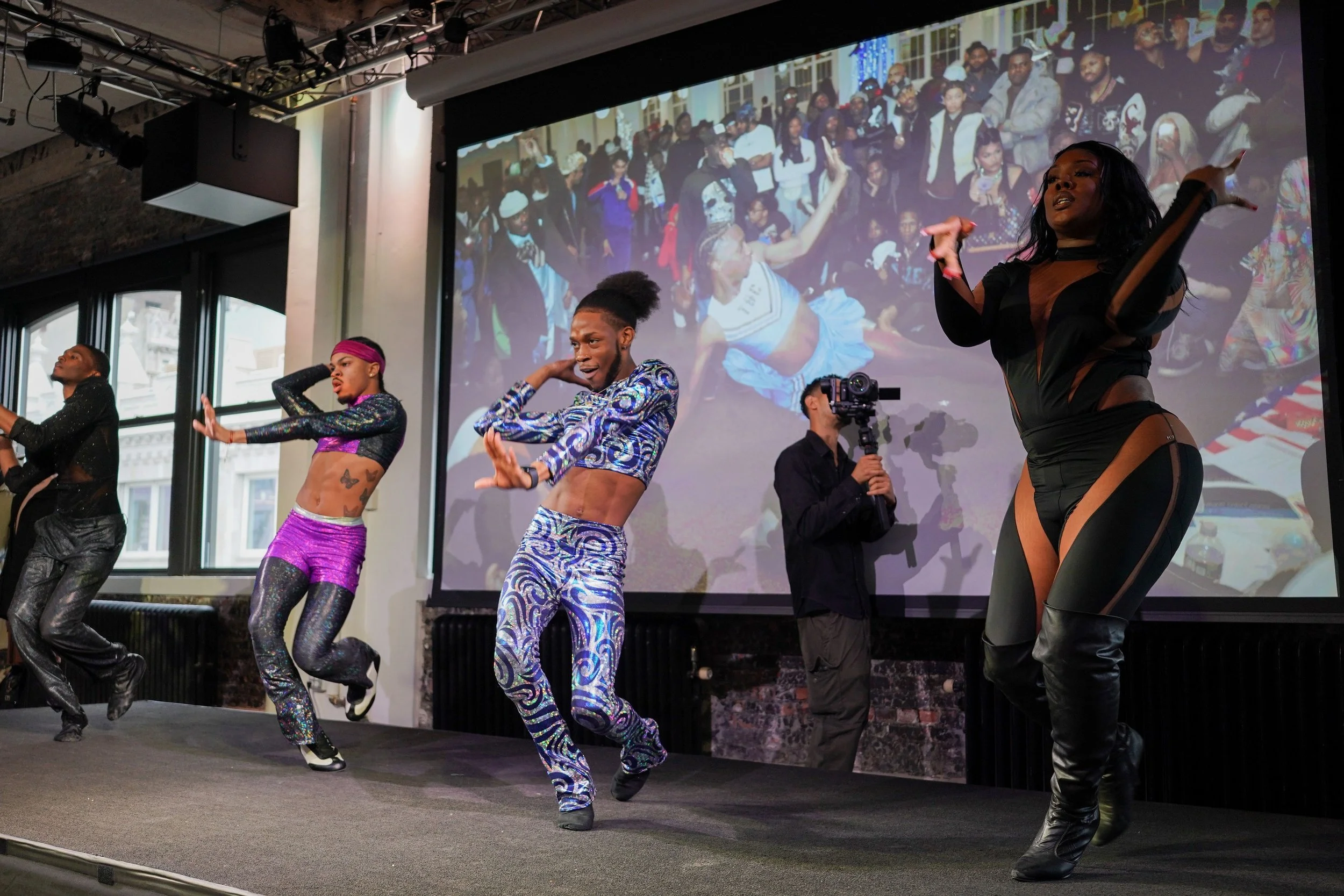A group of dancers performing on stage with an audience in the background on a large screen, featuring diverse performers in colorful outfits and energetic poses.