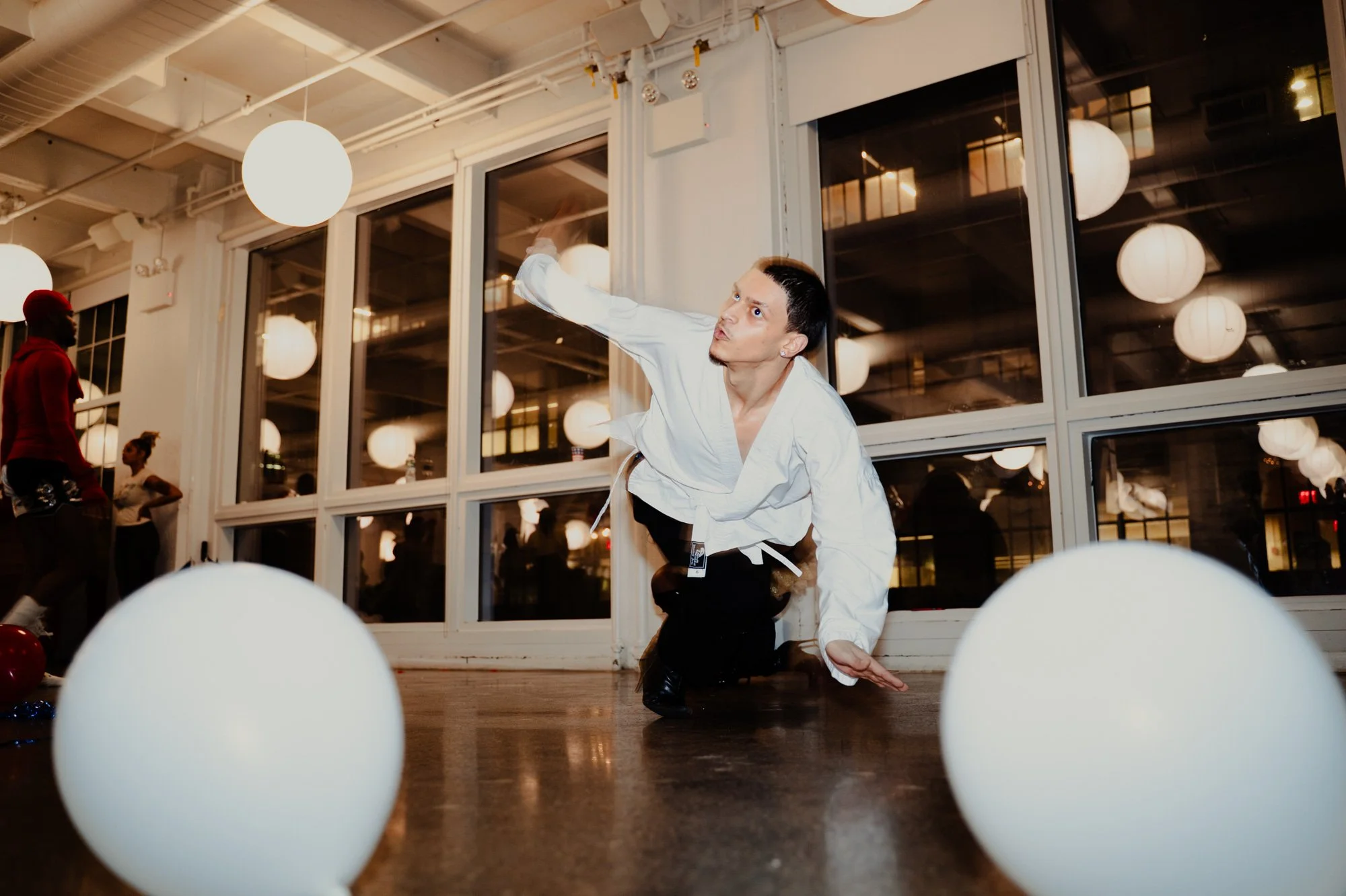 A man dressed in a white martial arts gi performing a move on the floor inside a room with large windows, round paper lantern lights, and balloons, while others watch in the background.