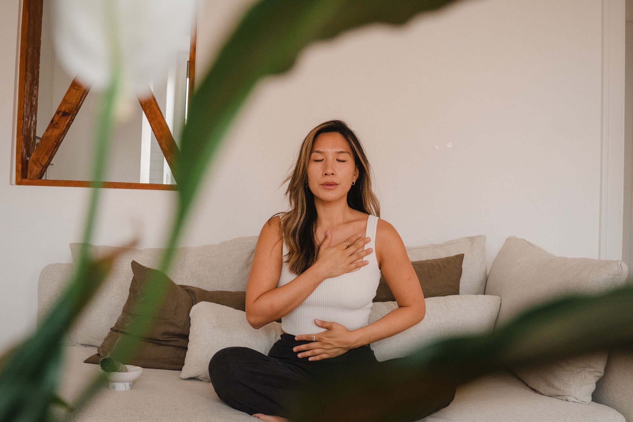 A woman practicing meditation at home, sitting cross-legged on a sofa with eyes closed, hand on chest and stomach, in a relaxed and peaceful state.