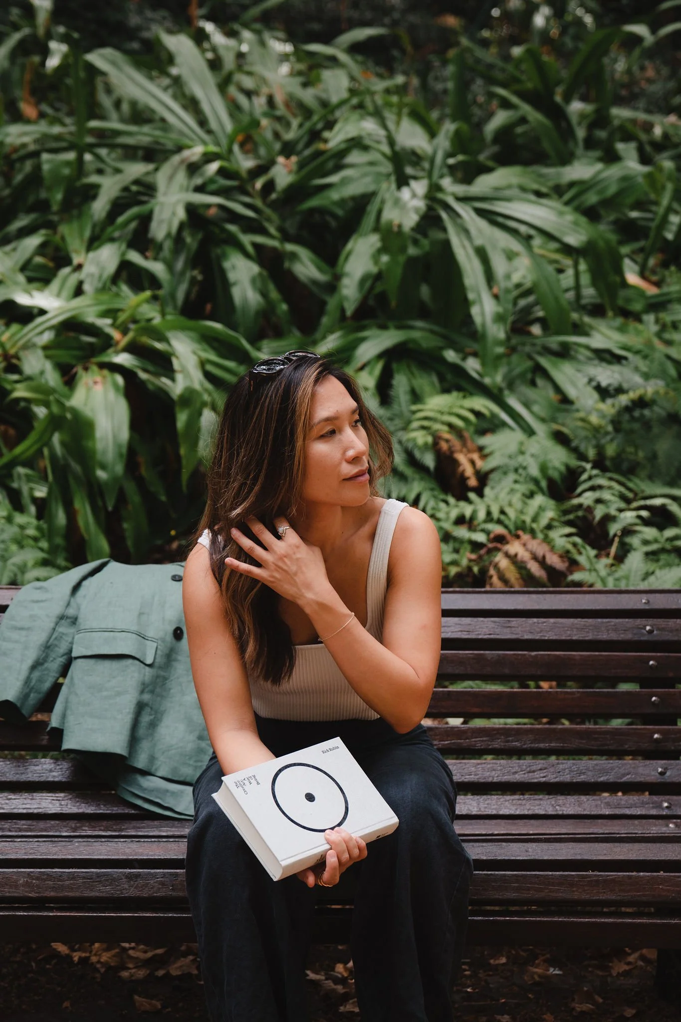 A woman sitting on a wooden park bench surrounded by green foliage, holding a book in her right hand and touching her hair with her left hand.